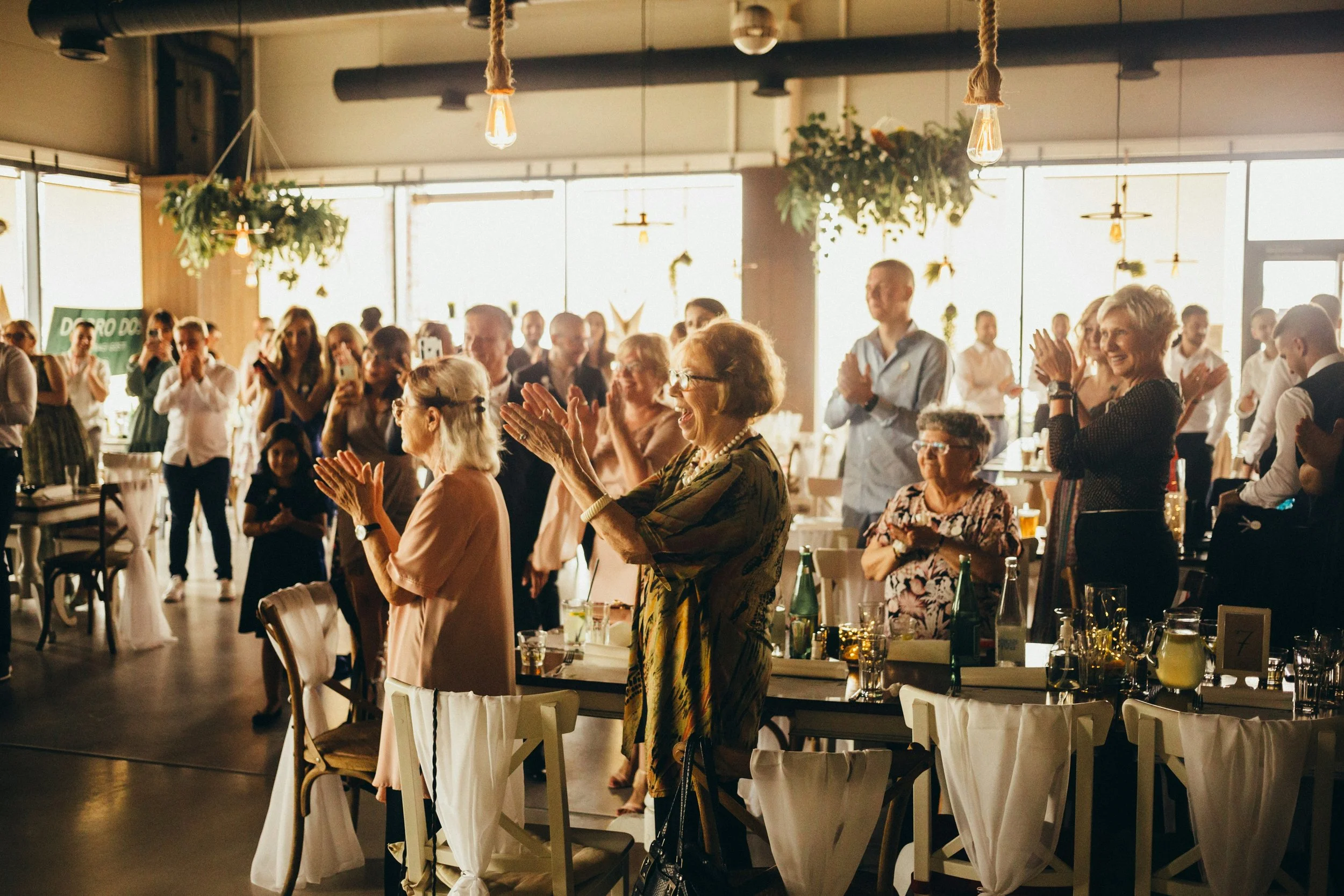 A group of people in a restaurant or event space are standing and clapping, smiling, and enjoying a moment of celebration or entertainment. The room has natural light coming from large windows and hanging decorative lighting.