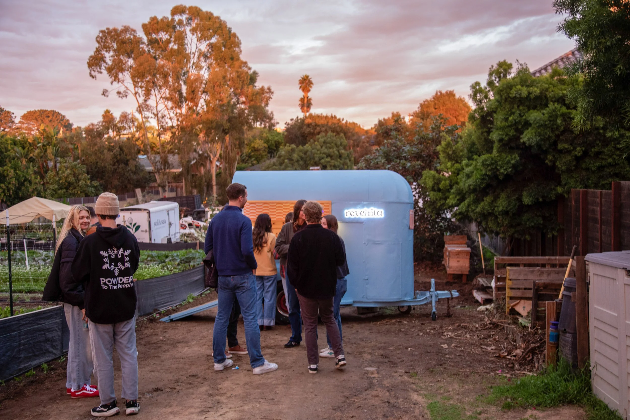 Group of people gathered outside a blue food truck named 'revelita' during sunset in a backyard with trees and gardening area.