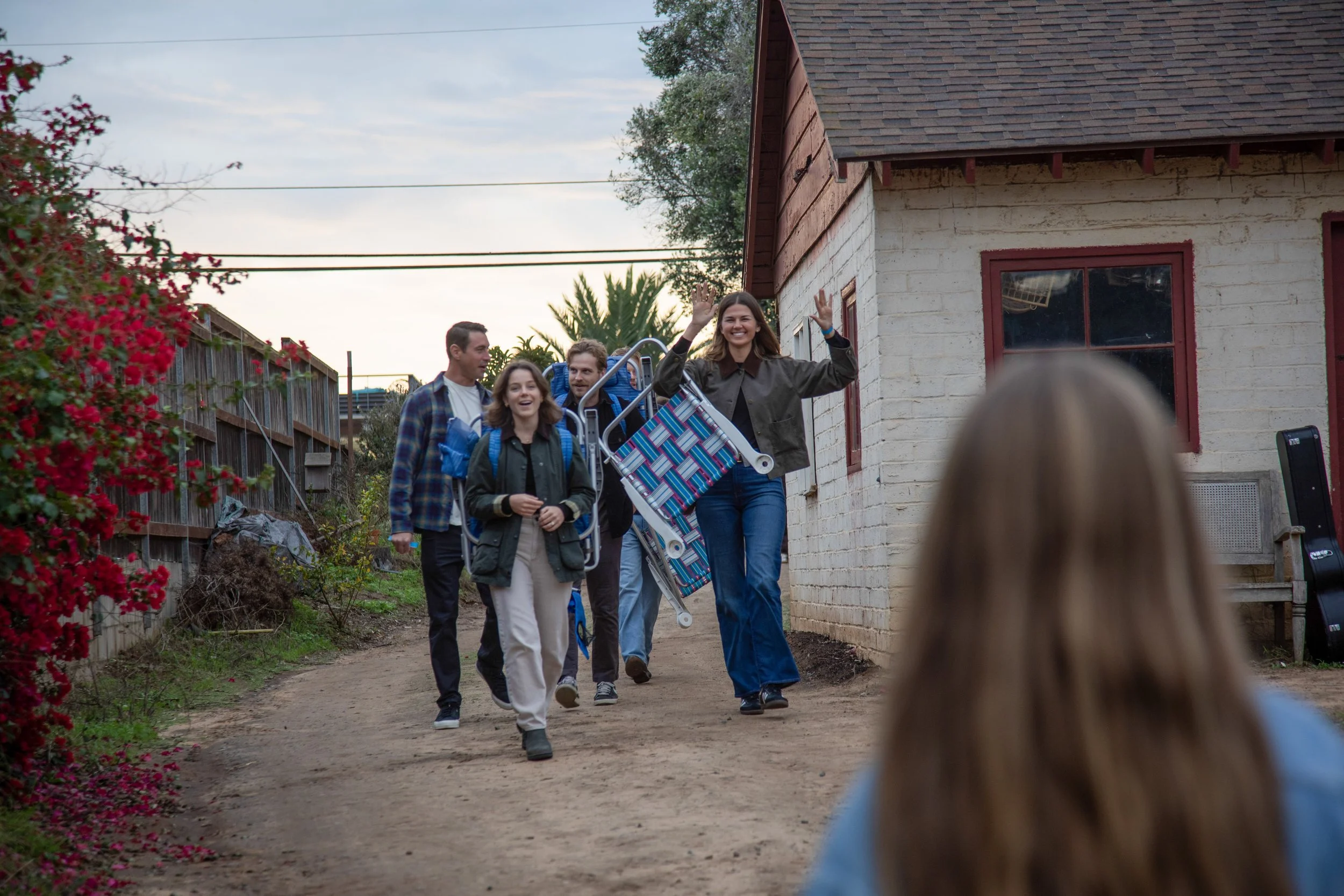 A group of five people walking on a dirt path outside a house, with one woman waving and smiling, carrying folded chairs, while another person in the foreground is taking a photo or filming.