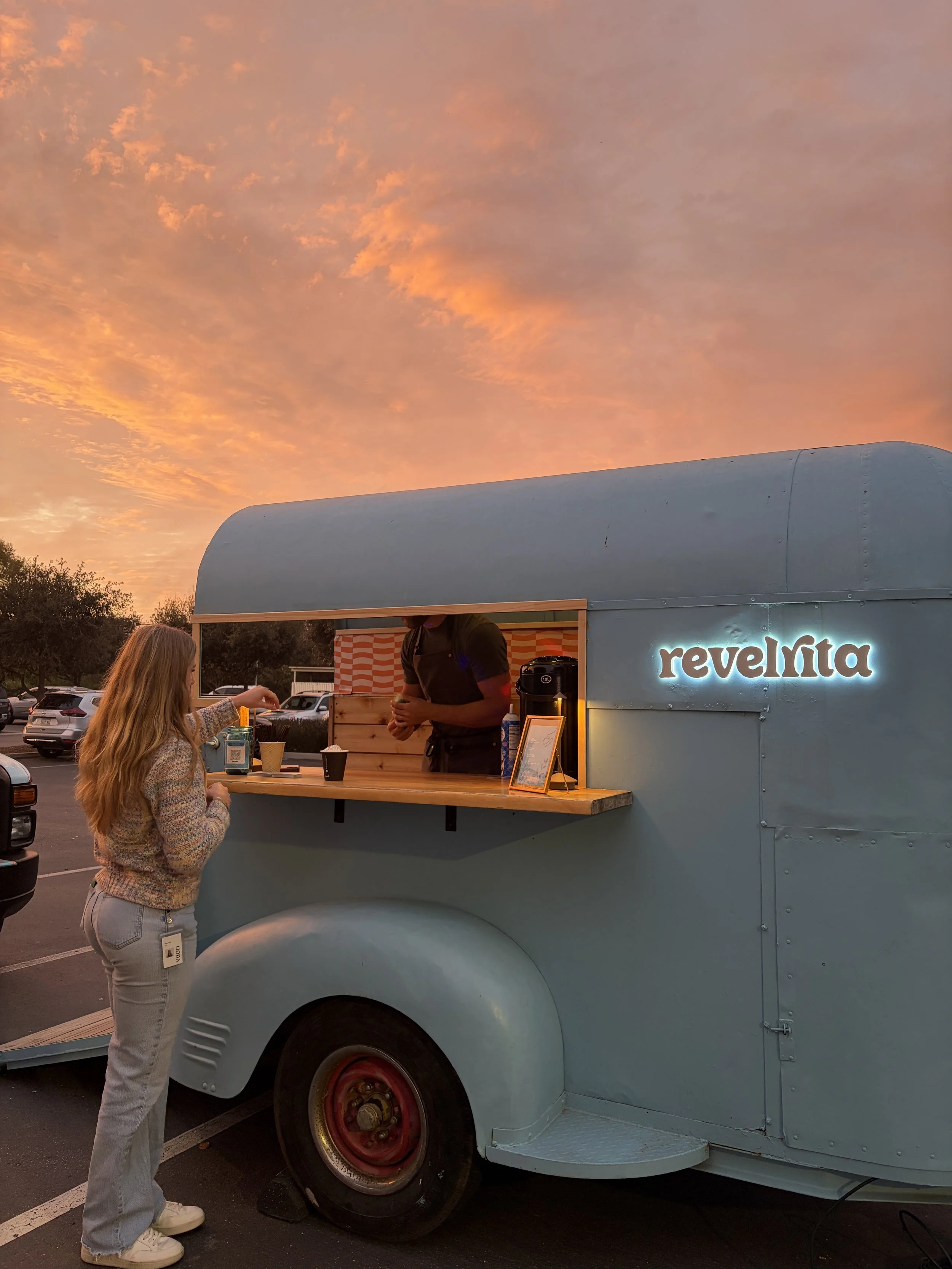 A woman ordering from a light blue food truck named 'Revelinta' during sunset, in a parking lot.