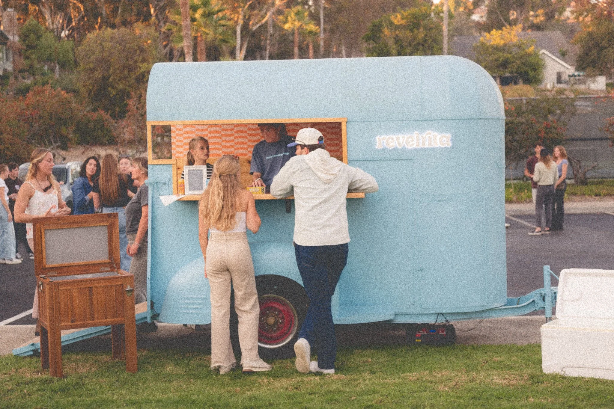 A light blue vintage food trailer with a service window, where two people are ordering and a third person is working inside. Two women are standing in front of the trailer, talking to each other. In the background, there is a group of people waiting in line and chatting in an outdoor parking lot with trees and houses visible.