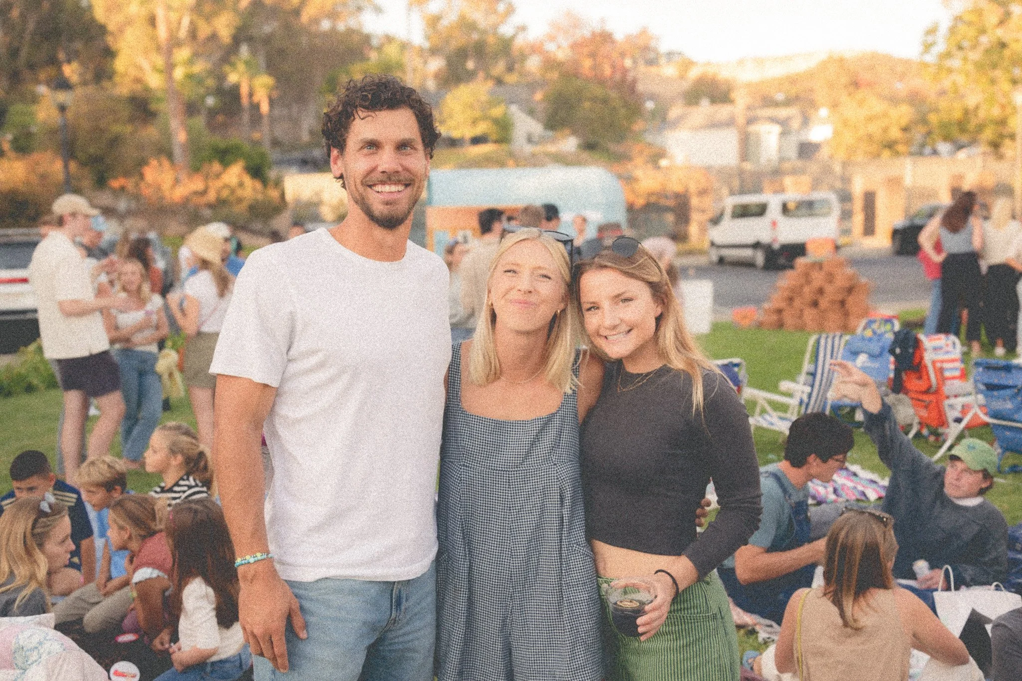 Three people smiling at a gathering outdoors during sunset, with additional people in the background sitting on the grass, talking, and enjoying food and drinks.