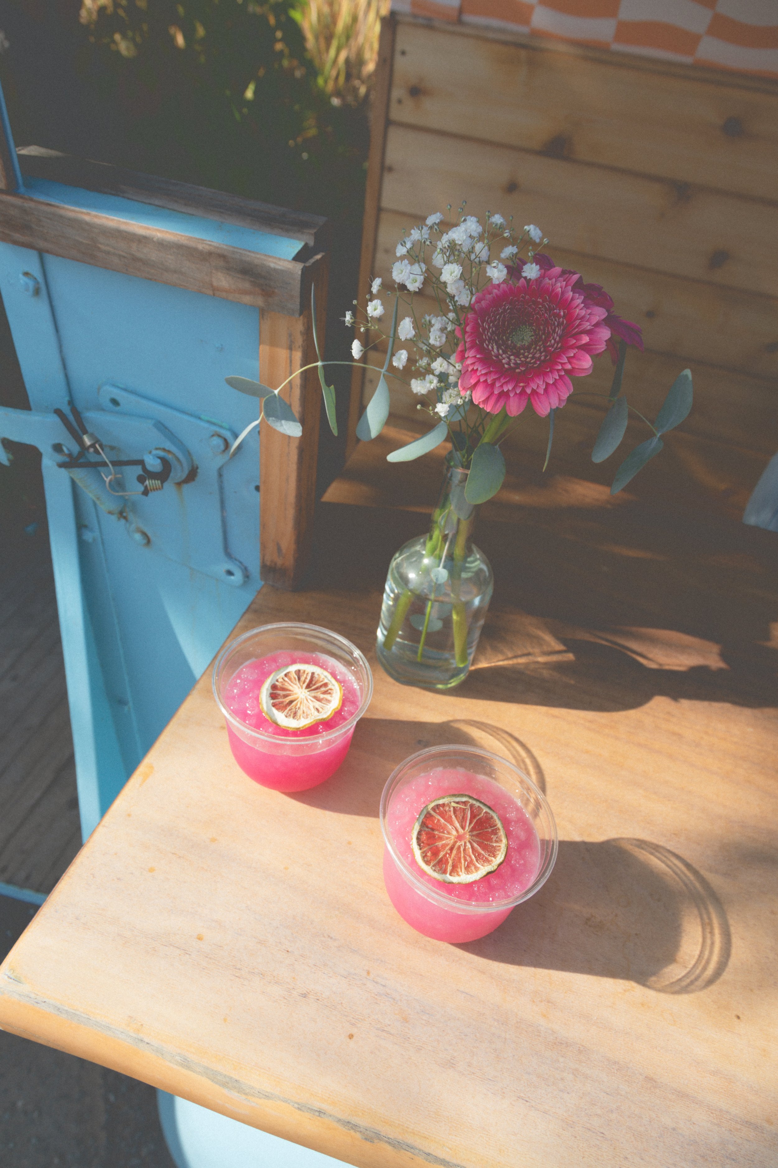 Two pink beverages with dried lime slices on top are placed on a wooden table, with a vase of pink and white flowers nearby, in an outdoor setting.