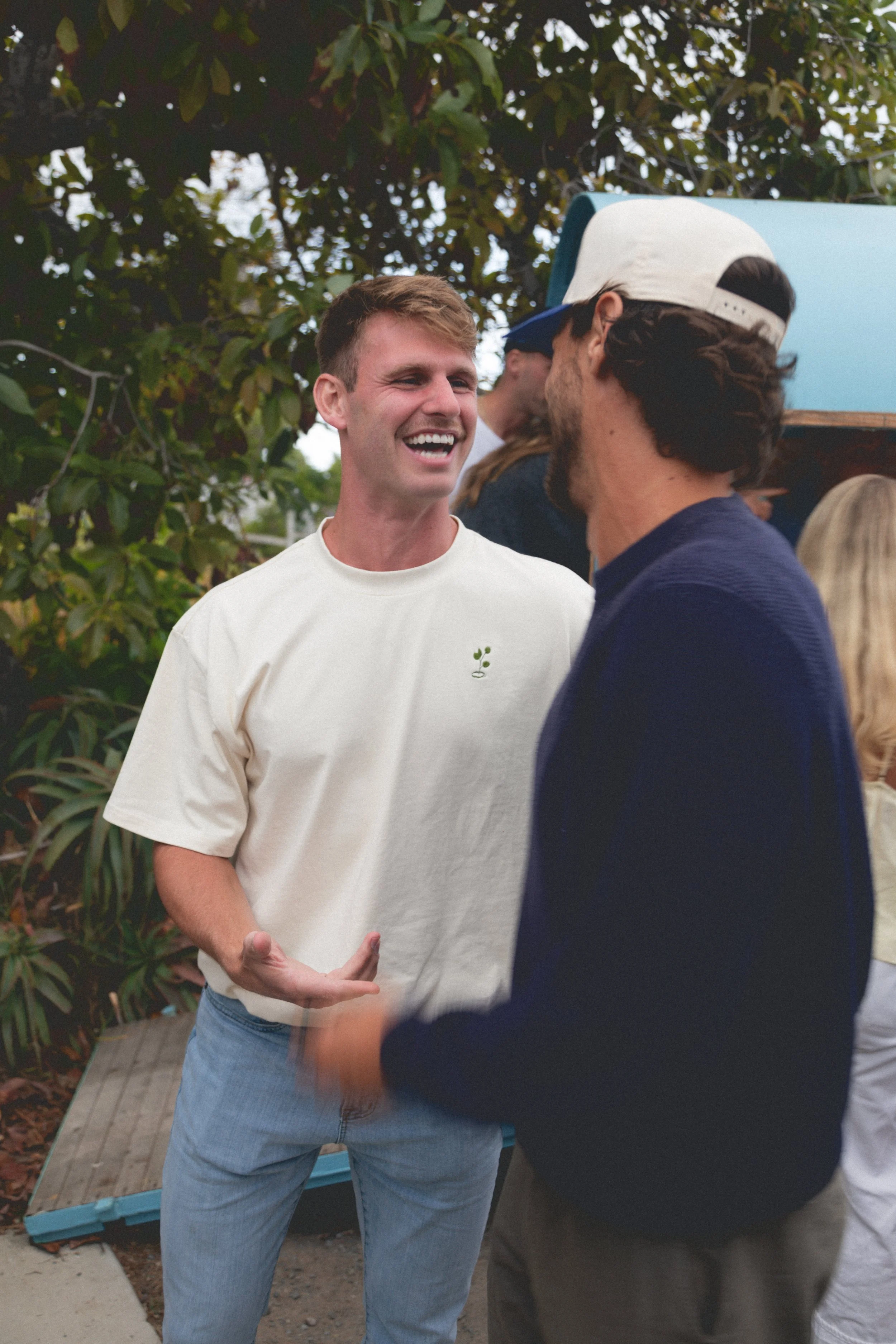 Two men are engaged in a conversation outdoors, smiling and laughing, with a group of people and greenery in the background.