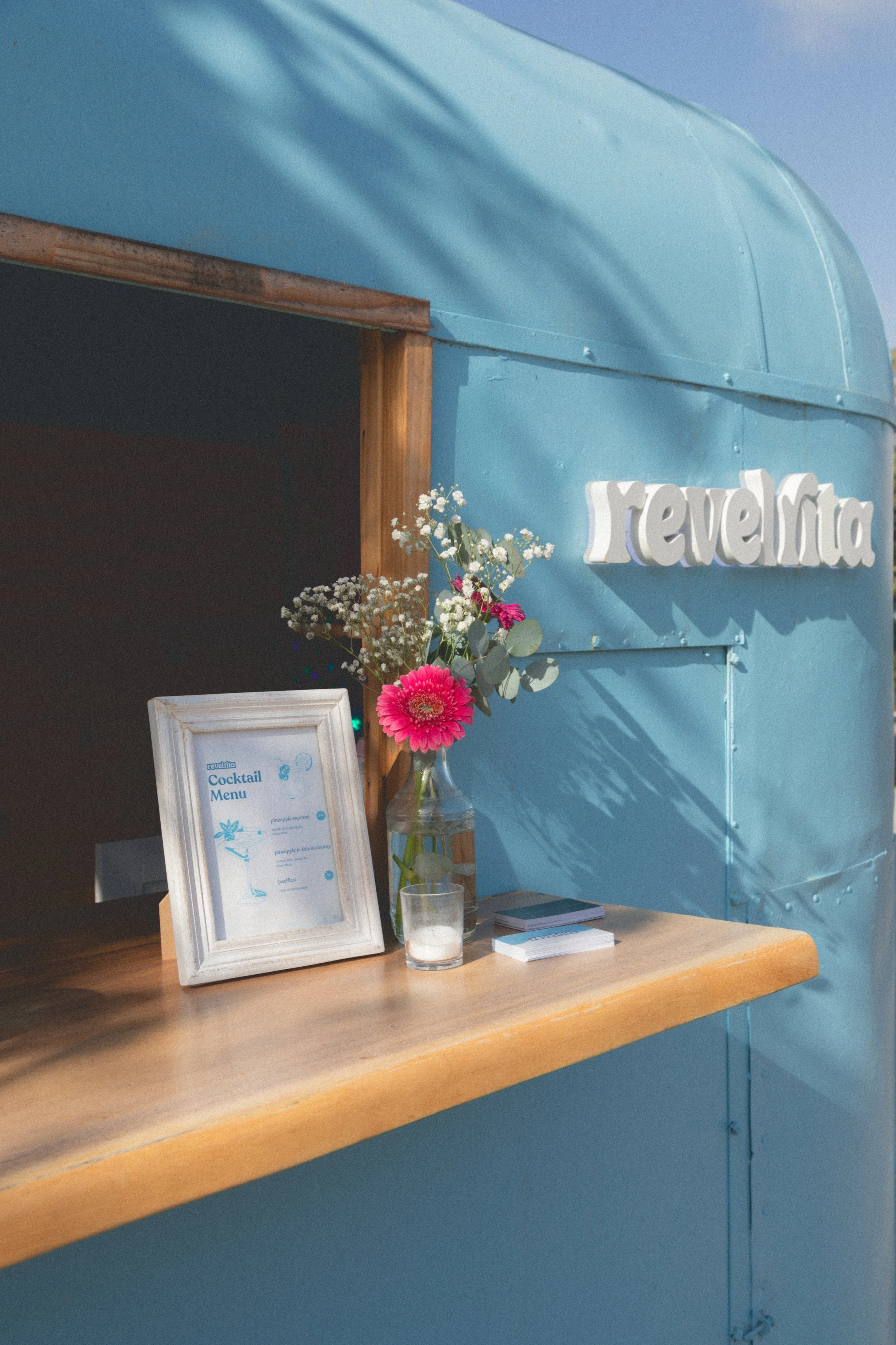 A wooden counter with a framed cocktail menu, a vase with pink and white flowers, and a small glass candle holder, against a light blue wall with the word 'revelio' in white 3D letters.
