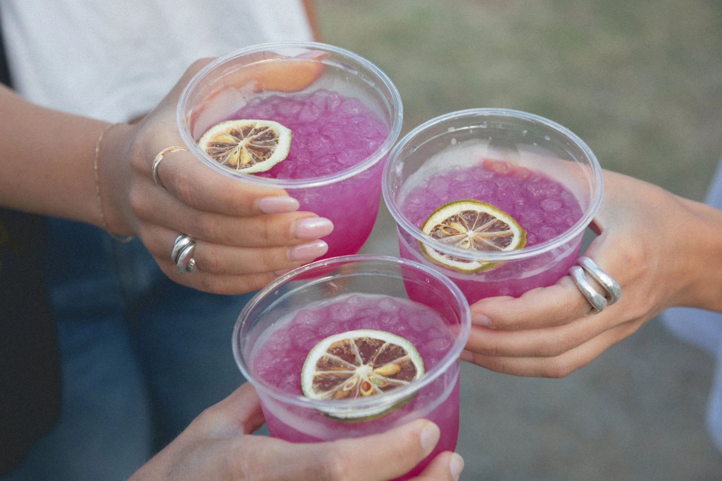 Three hands holding clear plastic cups filled with pink beverages and garnished with lemon slices.