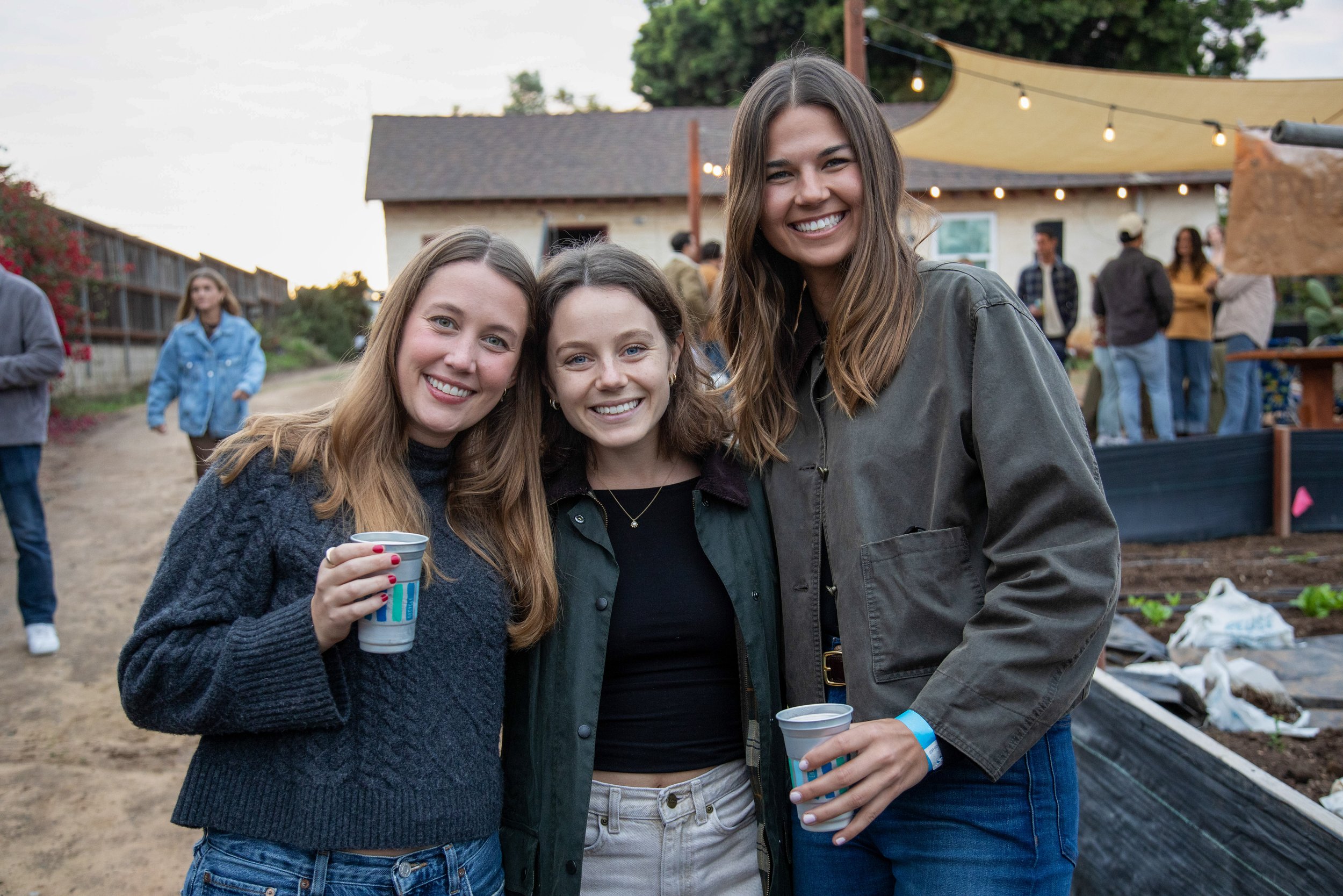 Three young women smiling and posing together at an outdoor gathering, holding drinks, with a group in the background near some tents and string lights.