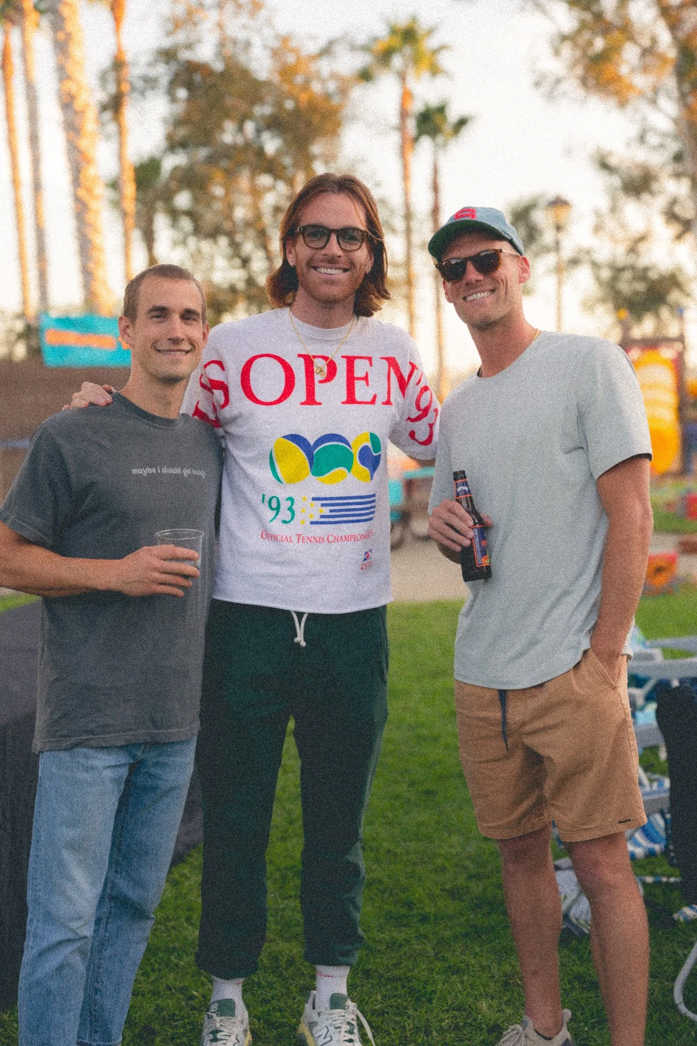 Three young men standing together outdoors during daytime, smiling, with trees and amusement park rides in the background. They are casually dressed, with two holding drinks.