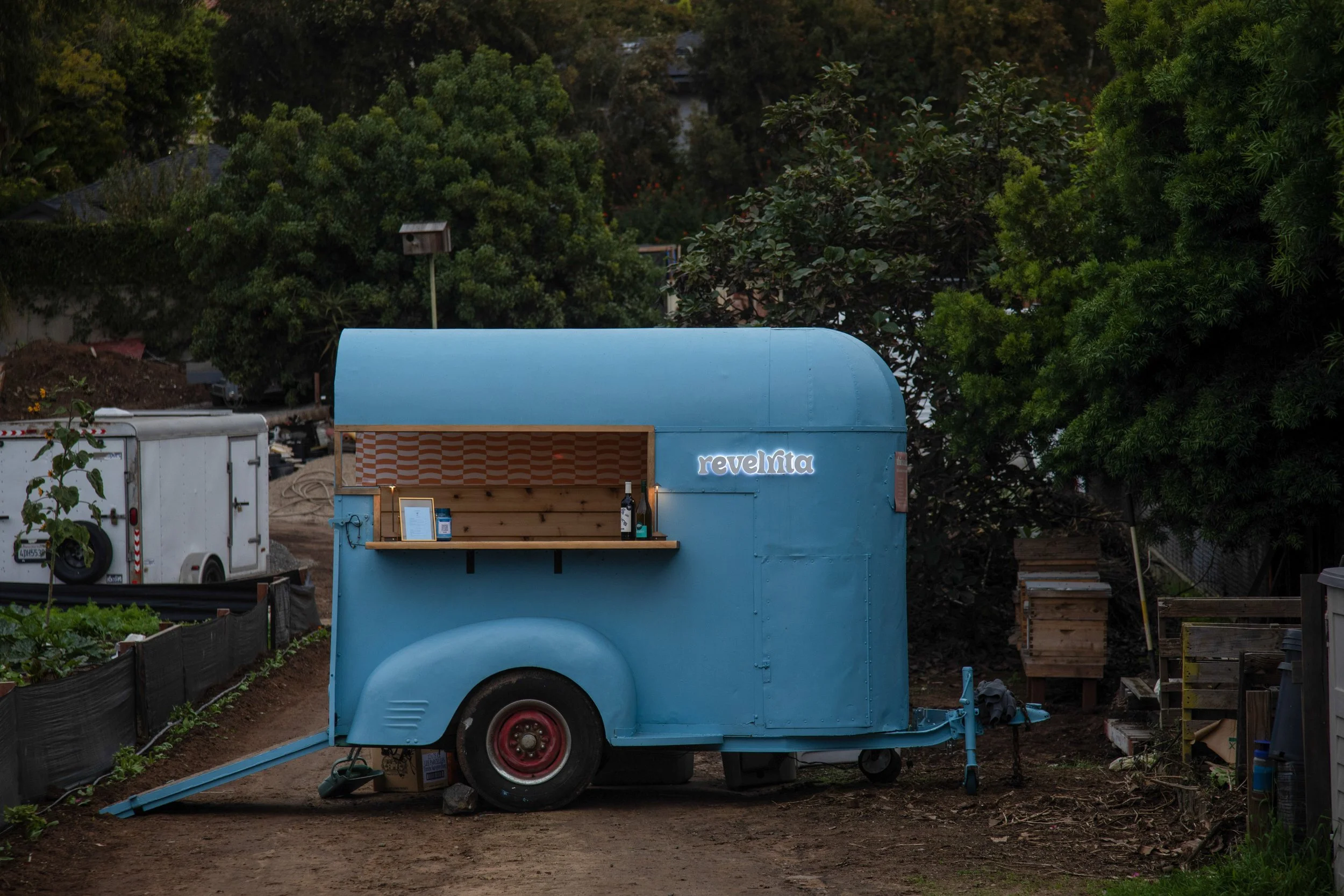 A small blue food trailer named 'revelata' parked on a dirt path, with a wooden shelf holding a bottle of wine and a framed menu, surrounded by greenery.