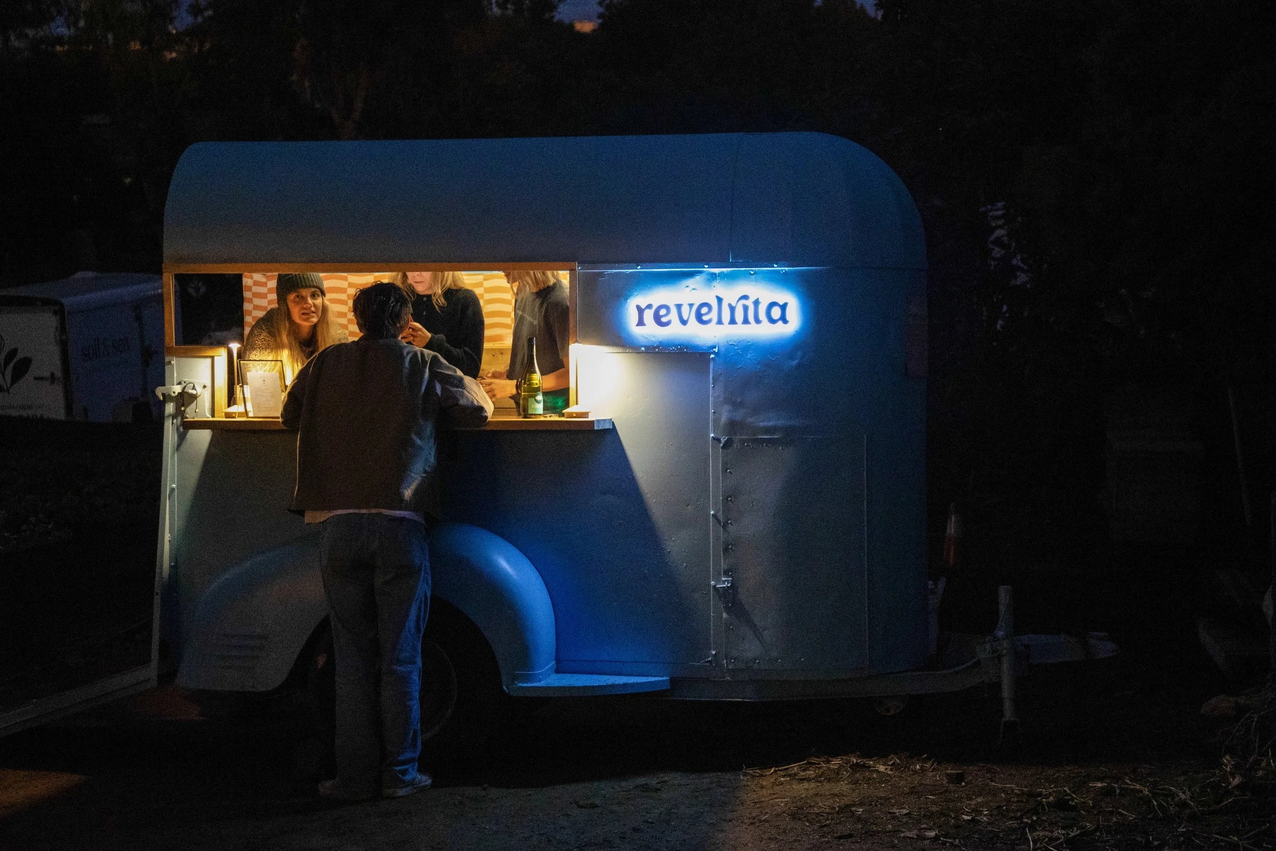 A person stands at a food truck counter at night, talking to three women serving inside. The truck has a bright neon sign that reads 'revelita'.