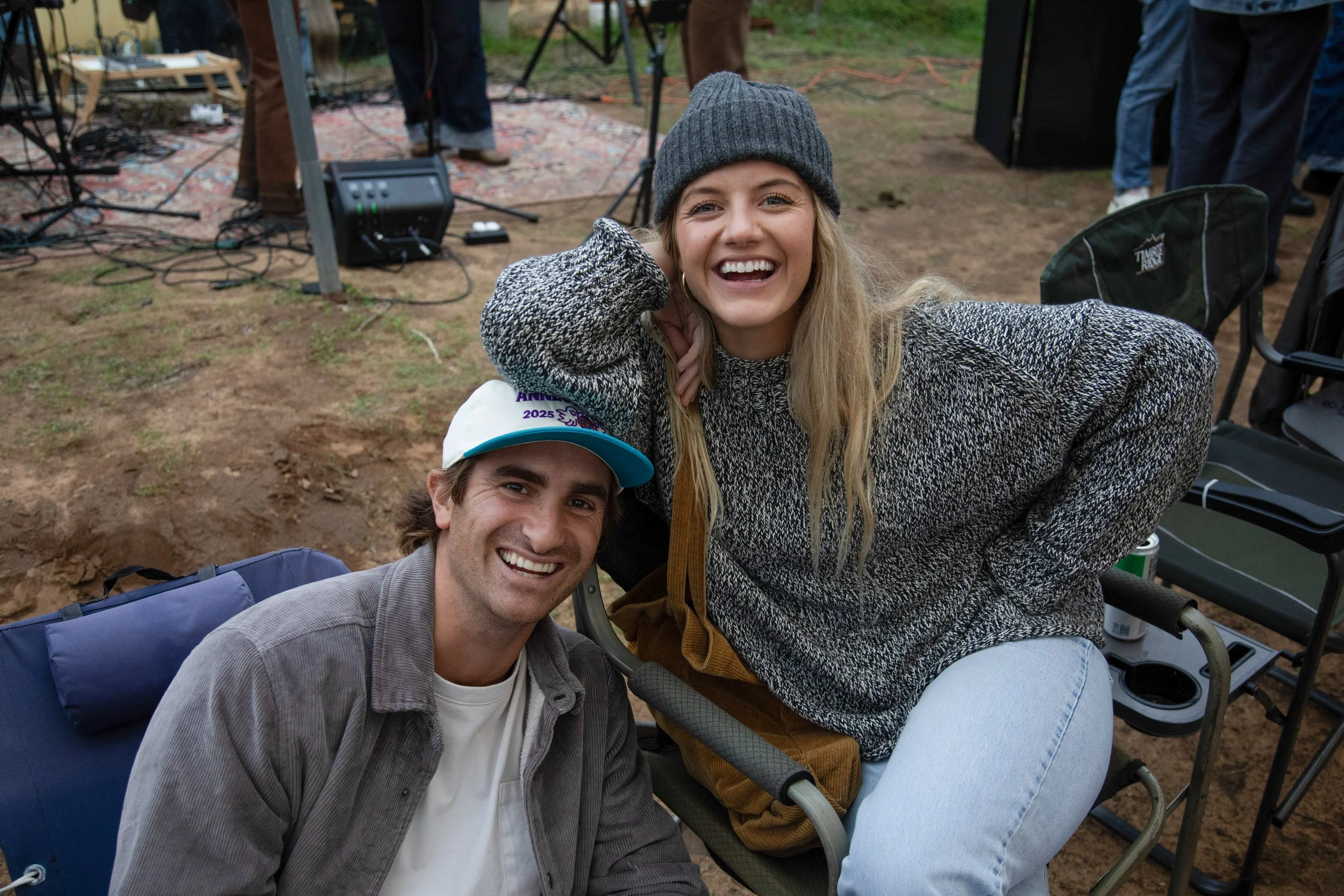 Two smiling people, a man and a woman, are outdoors at a gathering or event. The woman is wearing a gray beanie and a dark gray sweater, leaning on her hand, while the man is sitting, wearing a white baseball cap and a gray jacket. There are chairs, equipment, and a stage with musical instruments in the background.