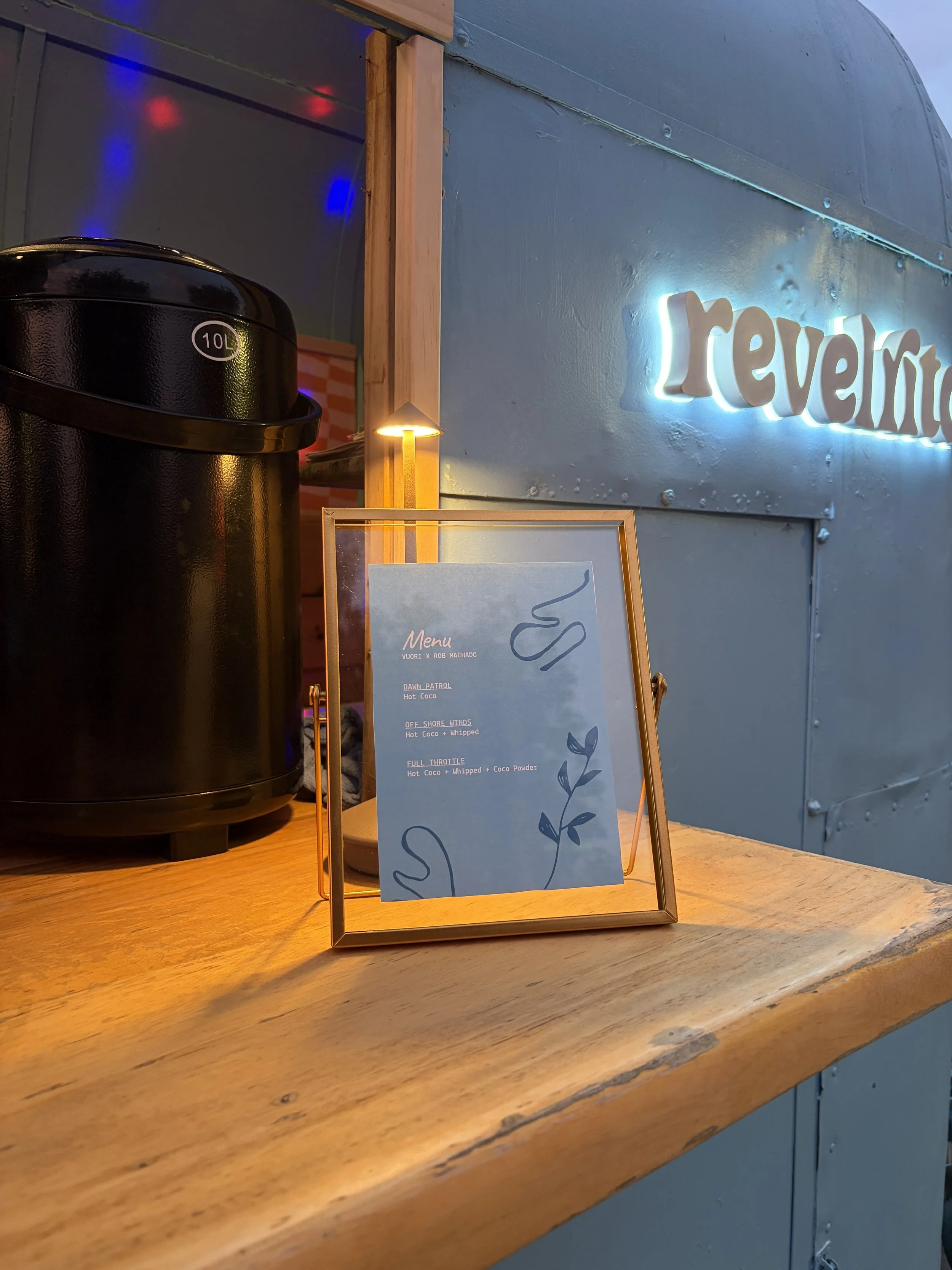 A framed menu stands on a wooden counter next to a black hot water dispenser. Behind, there is a light blue wall with illuminated "revol" signage and colorful lights.