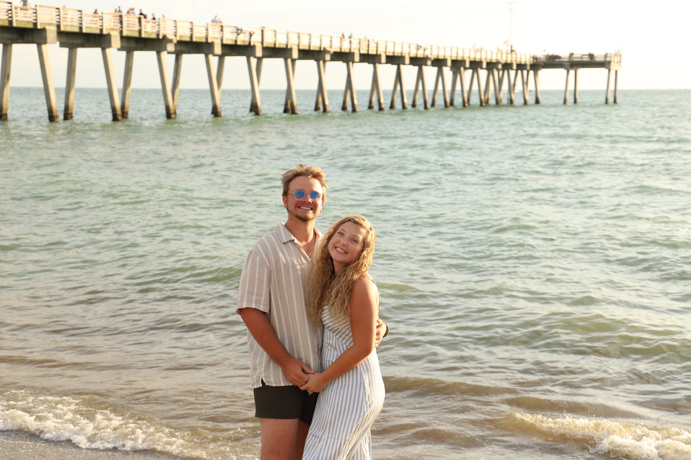 Smiling young couple standing on the shore at the beach with a pier in the background.