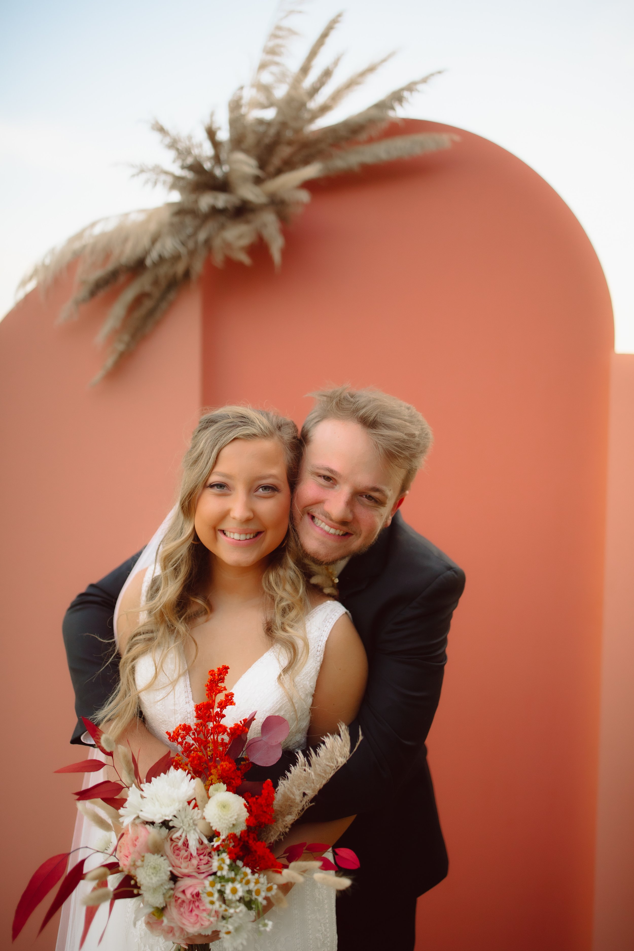 A happy bride and groom smiling and hugging each other, holding a bouquet of flowers, standing in front of a large pink and orange structure with pampas grass decorations.