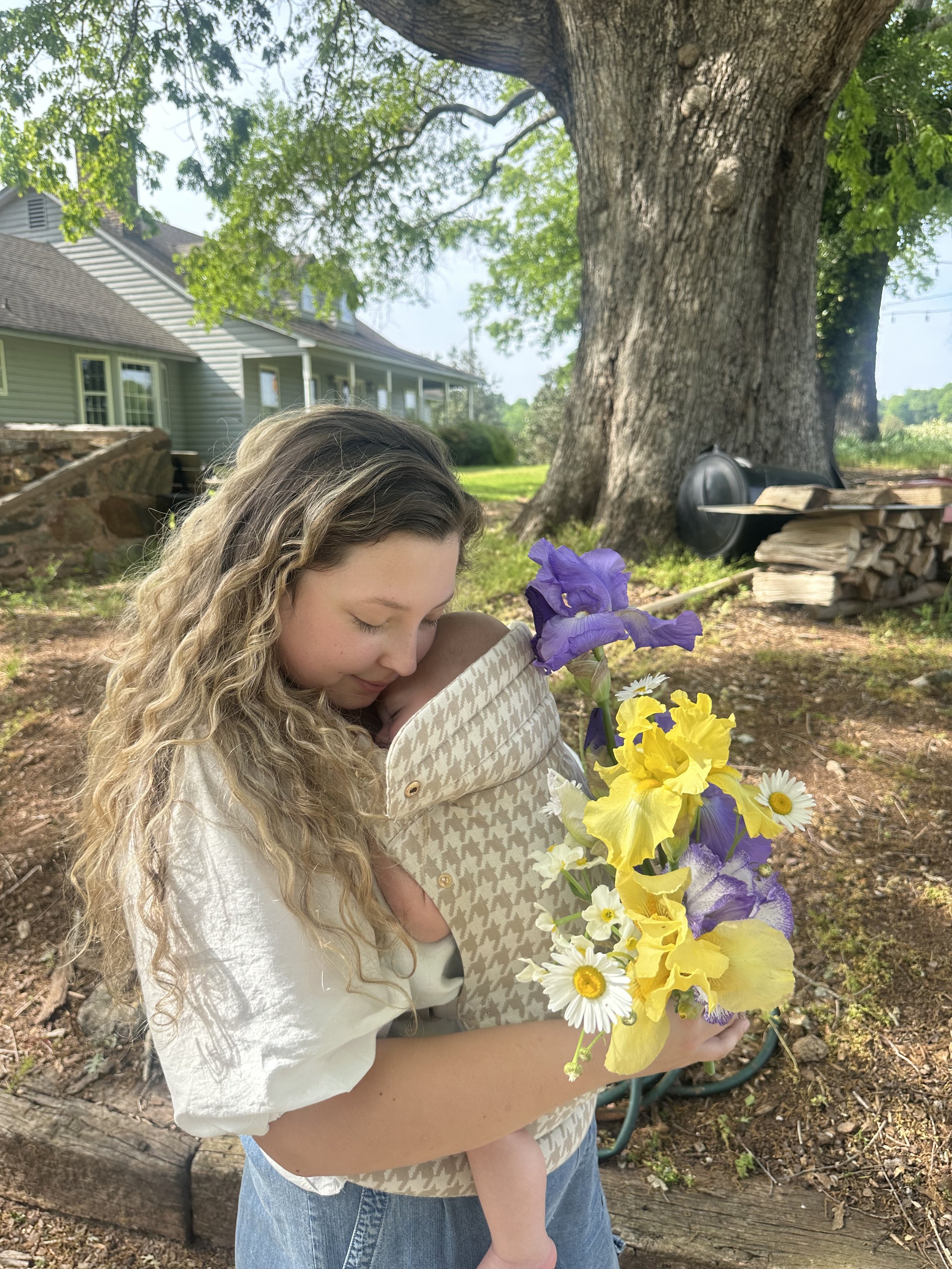 A woman holding a baby wrapped in a blanket with a houndstooth pattern, both outdoors under a large tree. The woman is gazing affectionately at the baby, and there is a bouquet of purple, yellow, and white flowers in front of them.