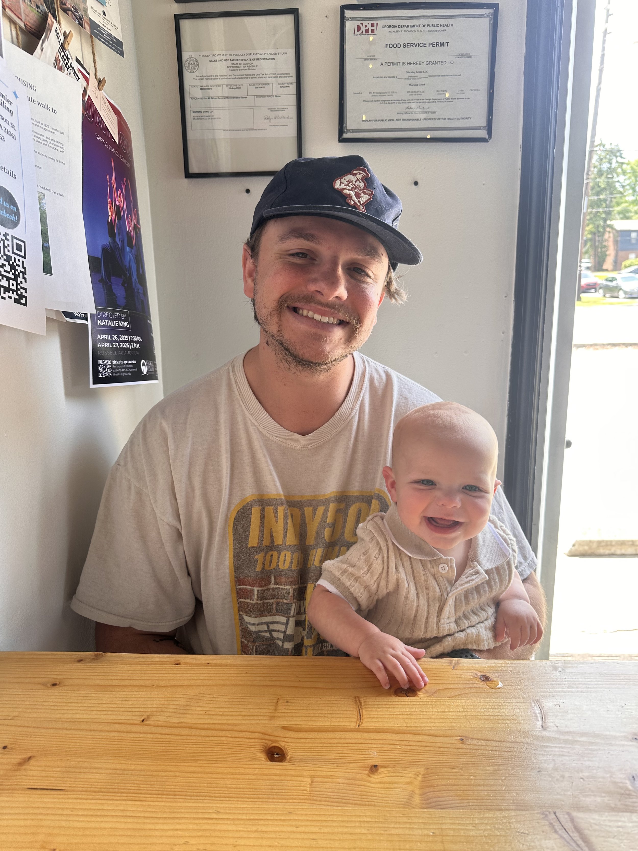 A man with a cap smiling while holding a baby girl with a big smile inside a restaurant or cafe, sitting at a wooden table near a door with sunlight coming in.