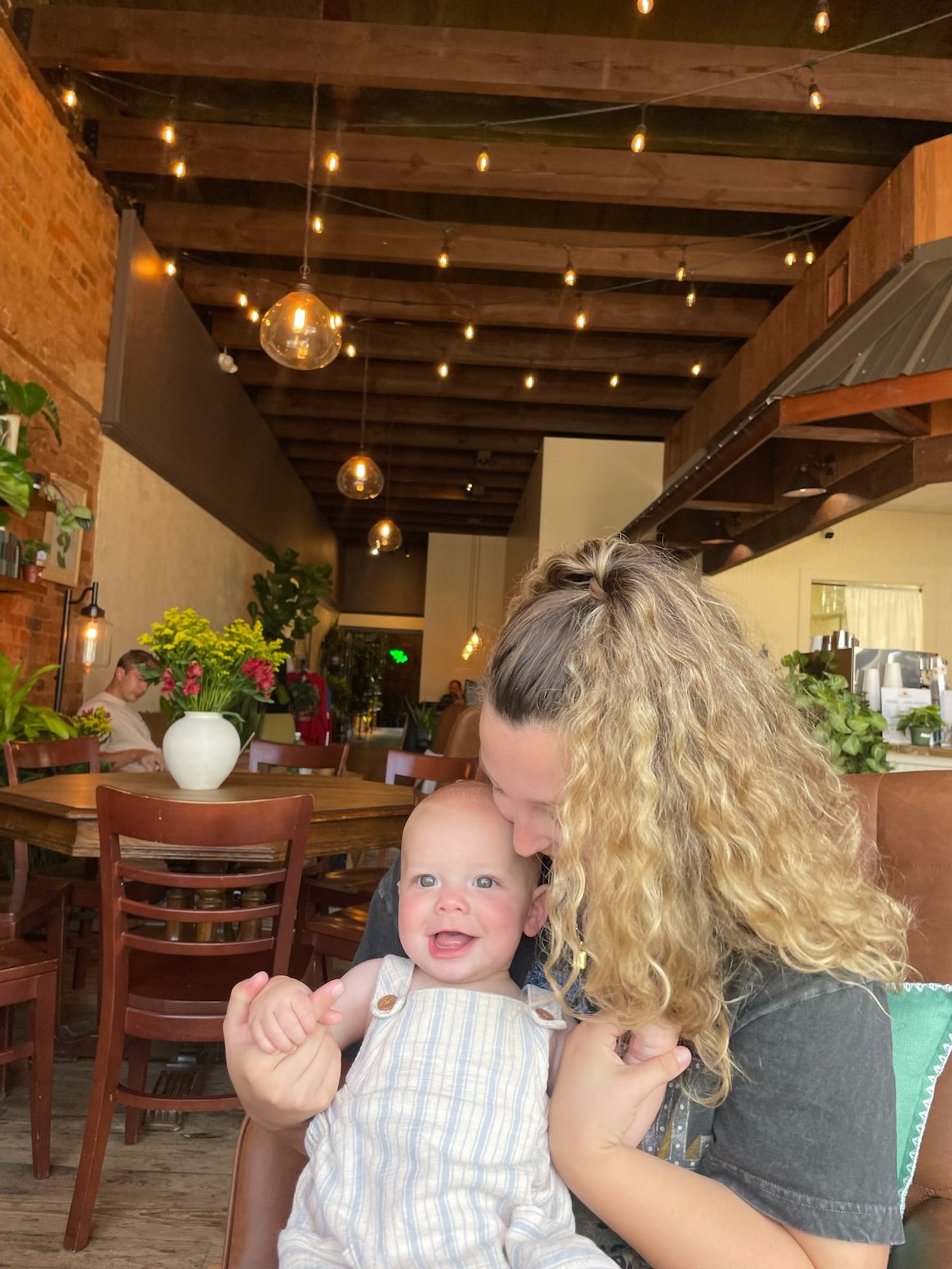 A woman with curly blonde hair kisses a happy baby with blonde hair in a restaurant. The baby is smiling and wearing a striped sleeveless outfit. The restaurant has warm lighting, wooden tables and chairs, and hanging string lights with exposed bulbs