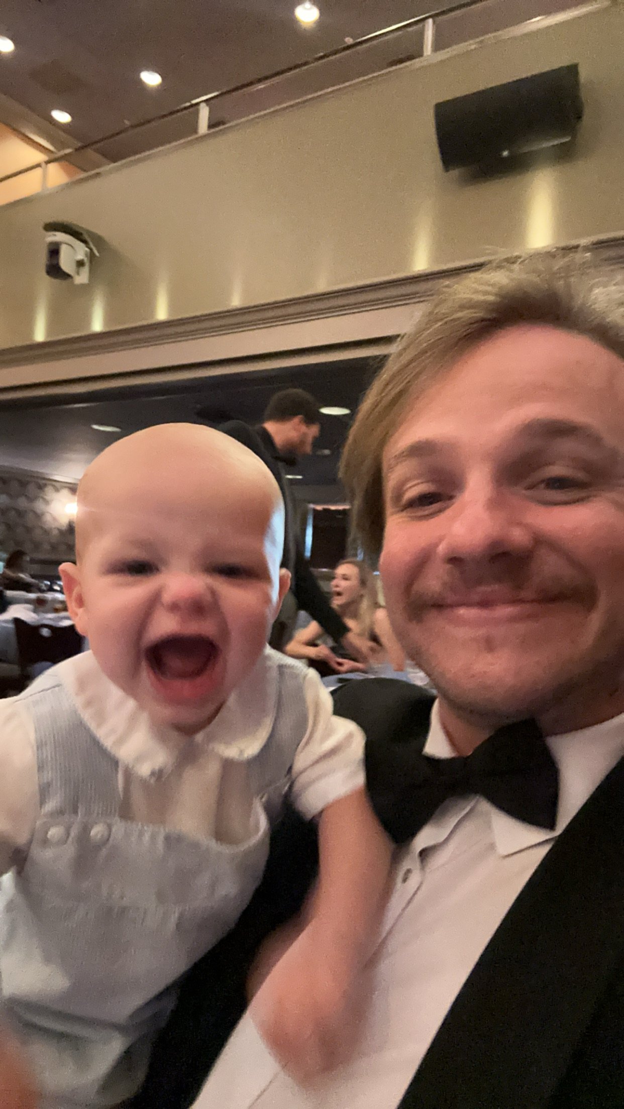 A man in a tuxedo taking a selfie with a laughing baby at a restaurant or event venue.