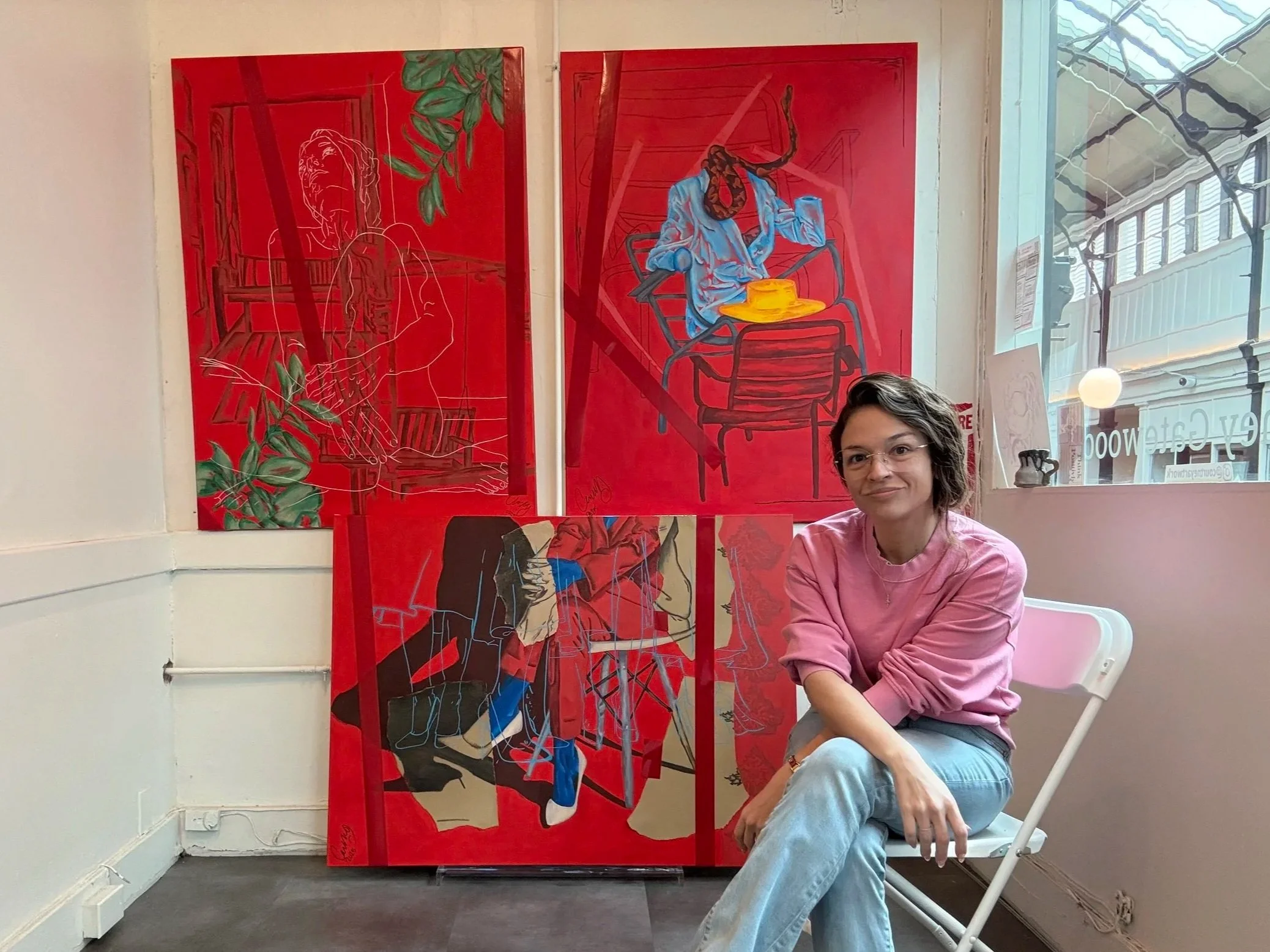 A woman in a pink sweatshirt and glasses sitting on a white chair next to four paintings on a wall inside a gallery or studio. The paintings feature abstract, colorful figures and scenes on red backgrounds, with some line art and paintings of seated figures.
