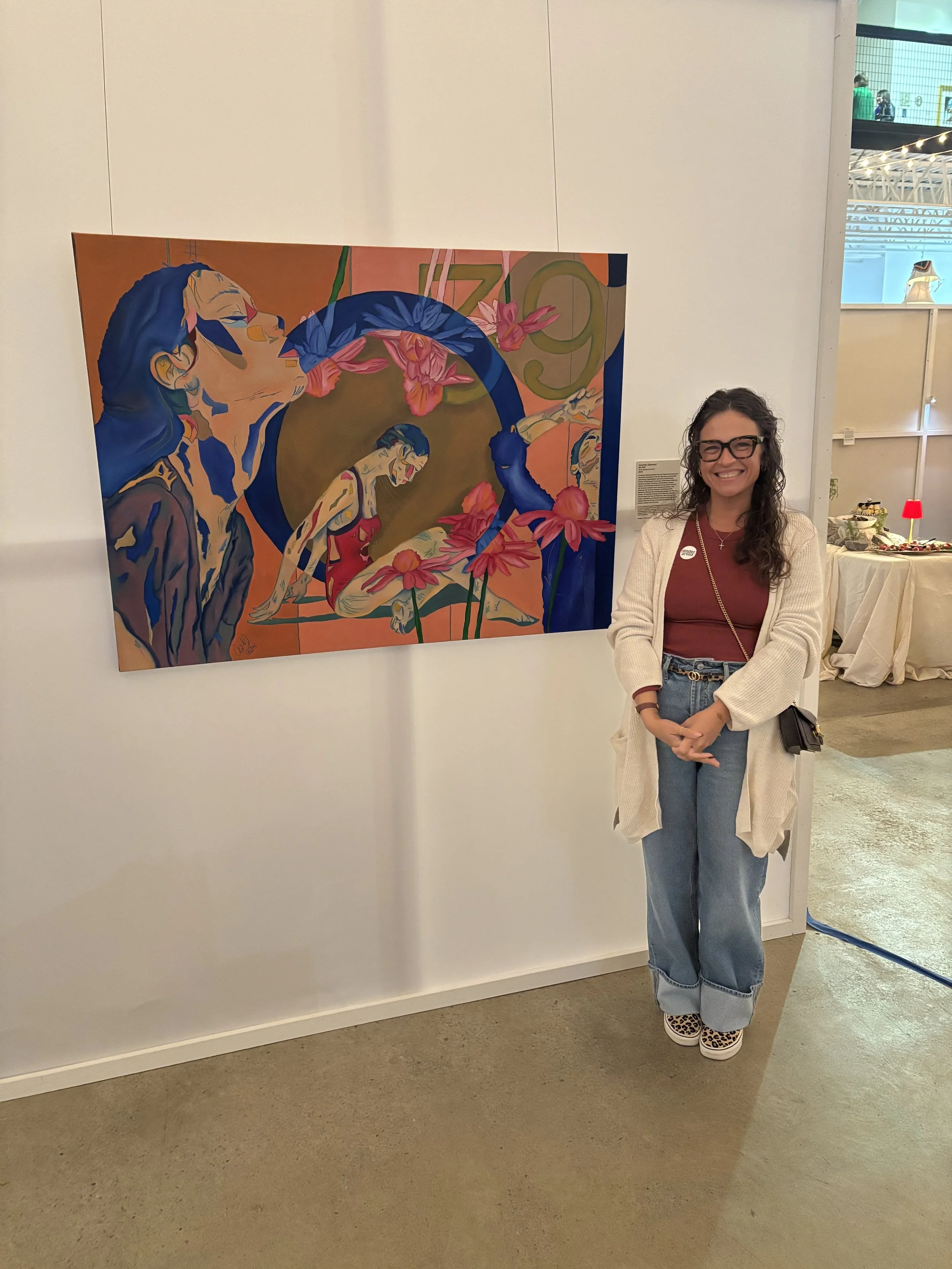 A woman with glasses smiling, standing next to a colorful abstract painting of a woman surrounded by pink flowers.