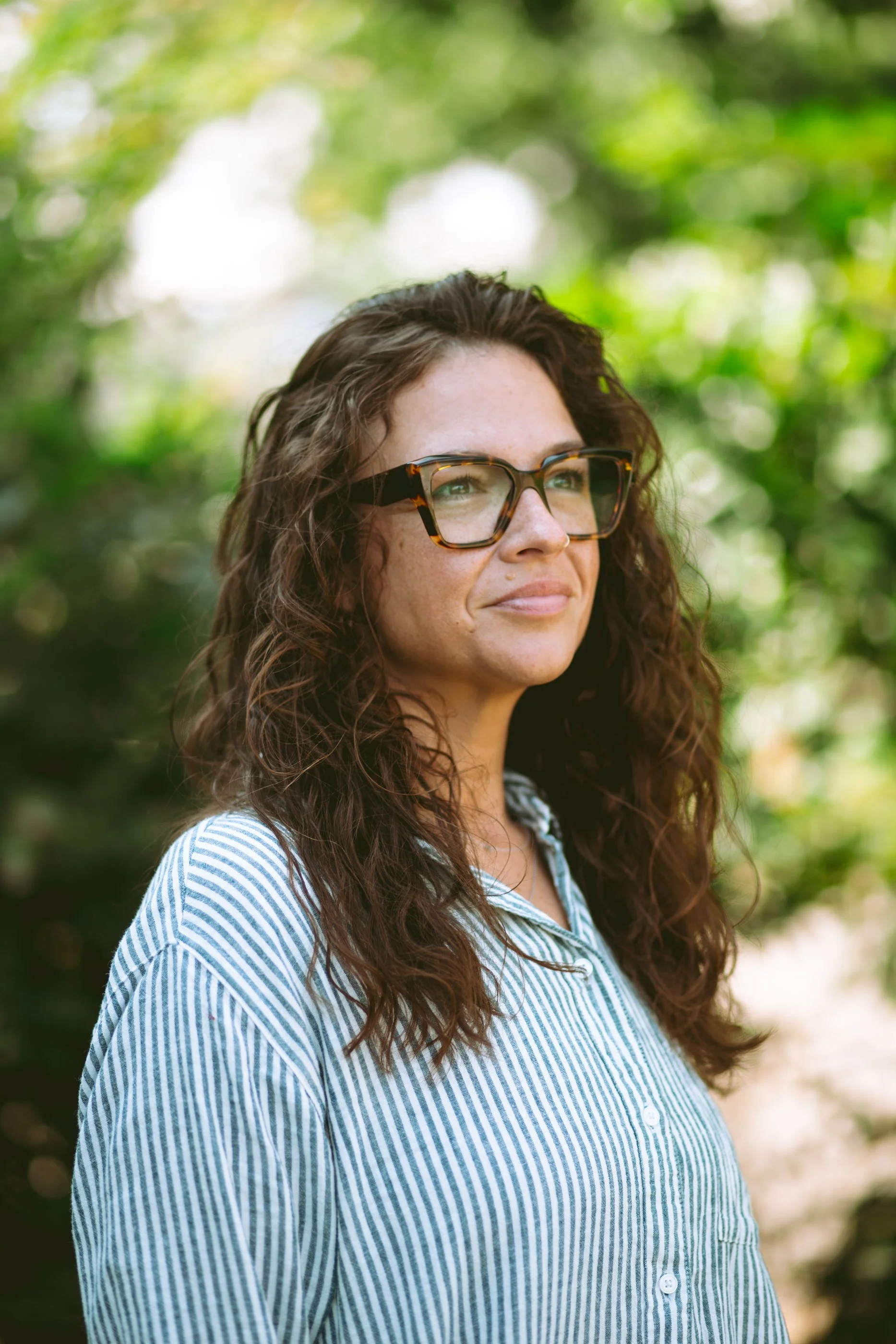 A woman with curly brown hair, wearing glasses and a blue and white striped shirt, standing outdoors with a blurred green background.