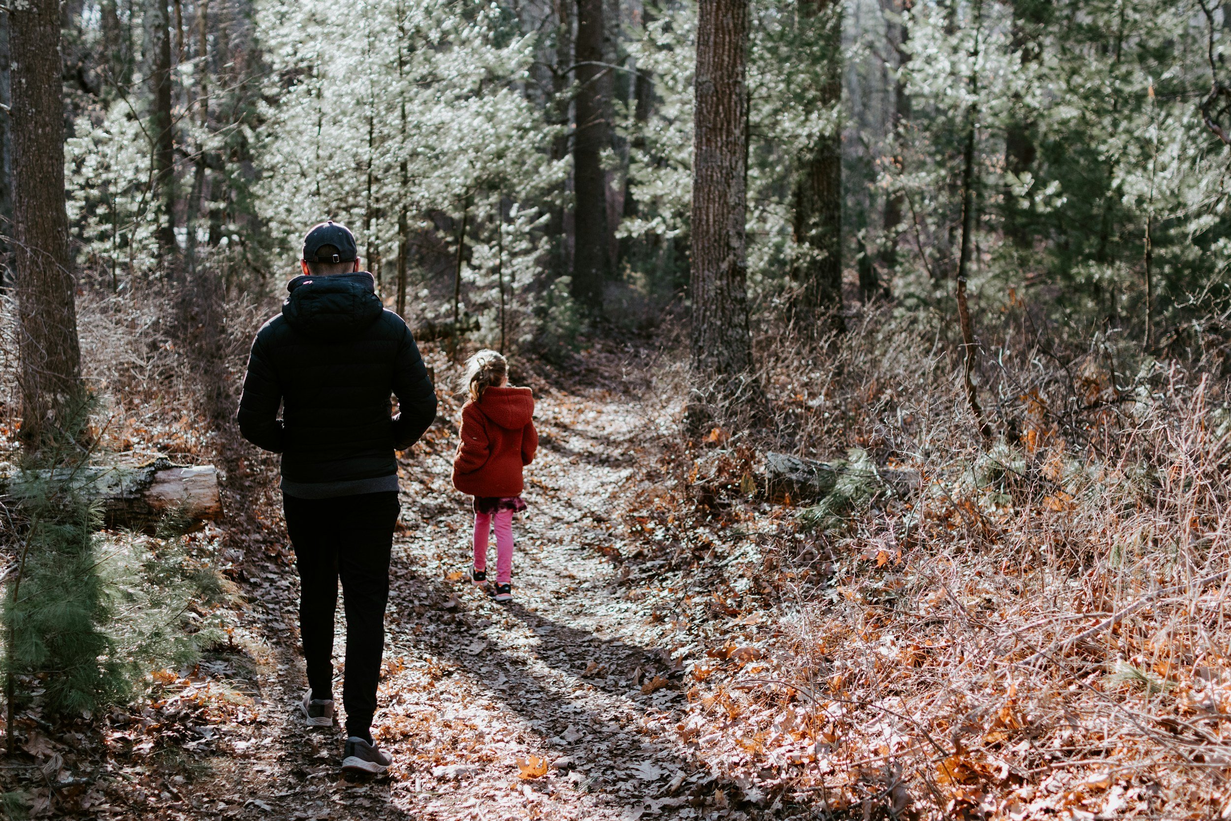 Dos personas caminando por un sendero en un bosque, rodeadas de árboles y hojas secas en el suelo.