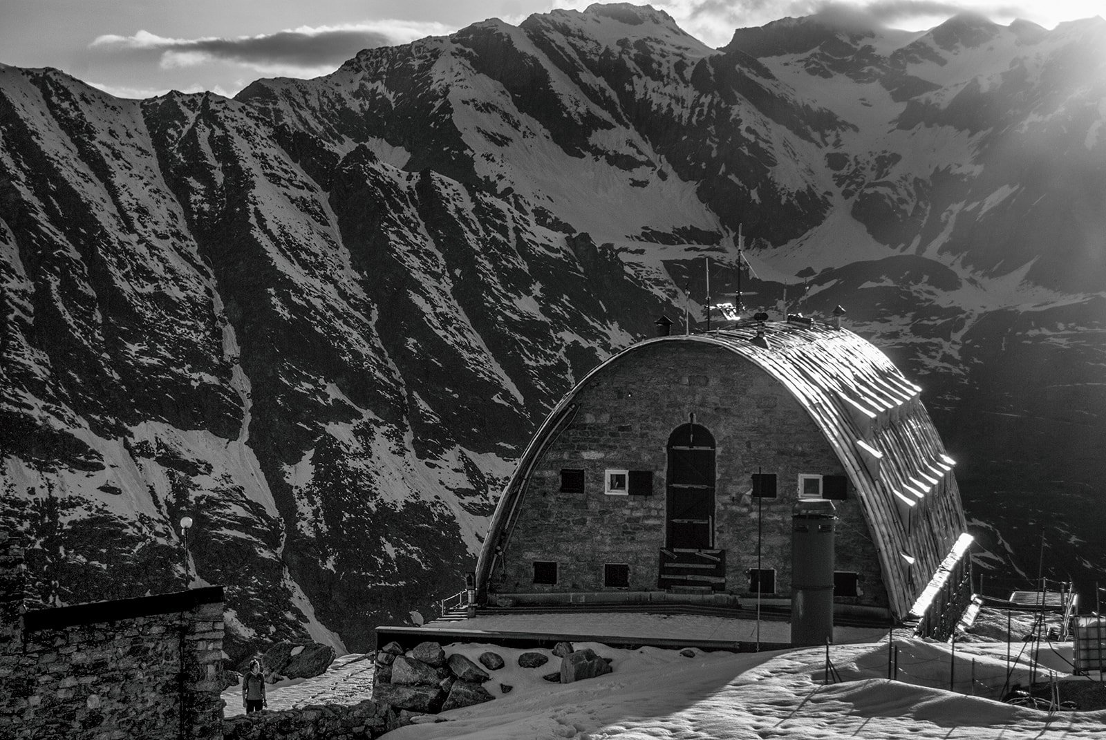 Refugio de montaña con techo curvado en un paisaje nevado, rodeado de altas montañas cubiertas de nieve, en blanco y negro.