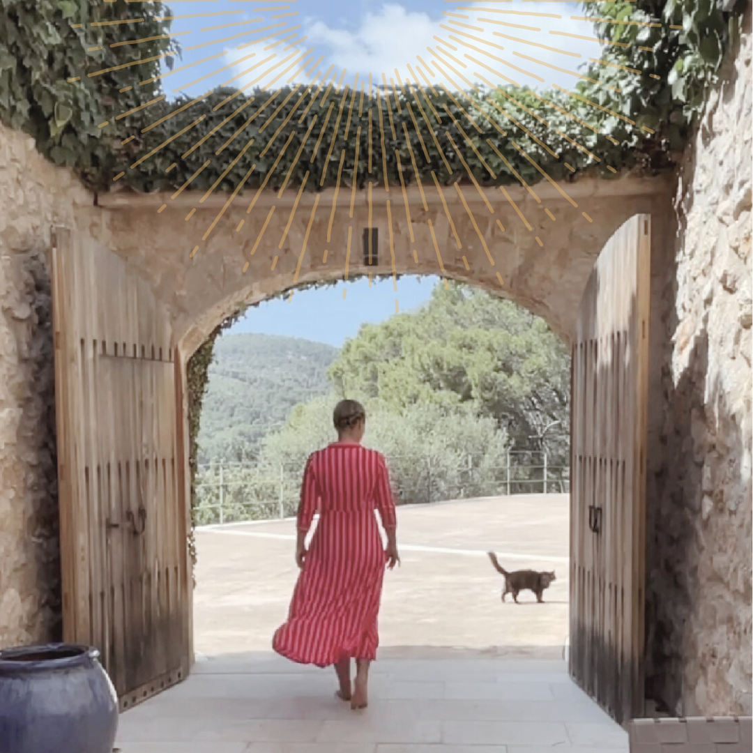 Eine Frau in einem roten, gestreiften Kleid geht barfuß durch eine offene Holztür, mit einer Katze im Freien auf einer Terrasse und grünem Baum im Hintergrund.
