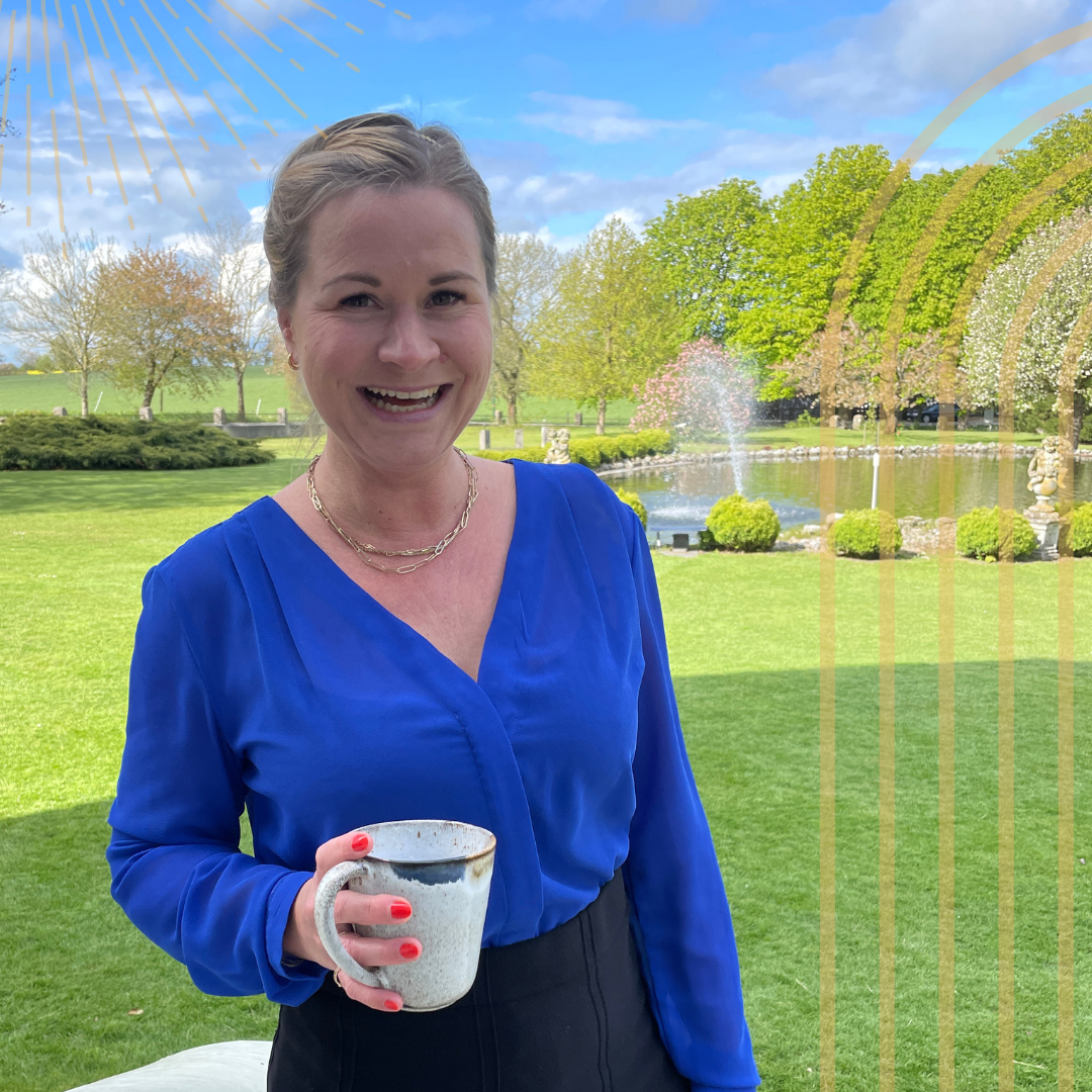 Eine lachende Frau mit kurzen hellen Haaren trägt eine blaue Bluse und hält eine Kaffeetasse in der Hand. Sie steht im Freien vor einer grünen Wiese mit Bäumen, einem Springbrunnen im Teich und einem blauen Himmel mit einigen Wolken.