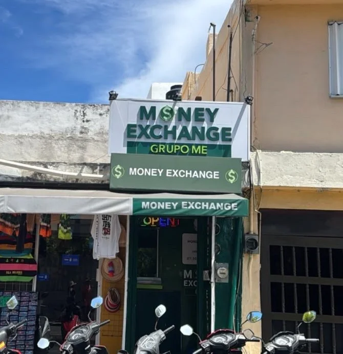 Tienda de cambio de dinero llamada 'Money Exchange' con un cartel que dice 'Grupo ME' en la parte superior, en una calle con paredes amarillas y cielo azul.
