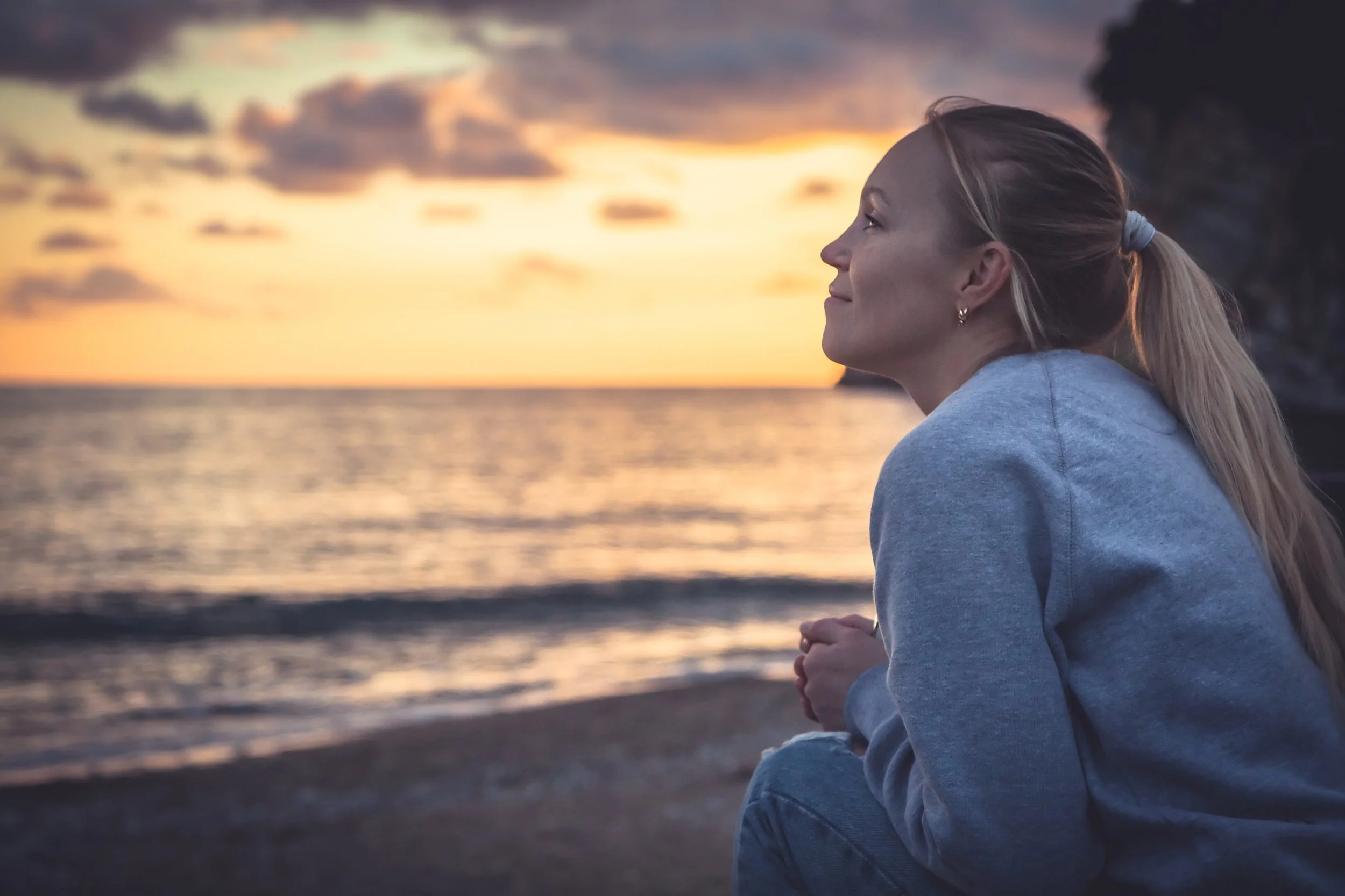 A woman sitting on the beach at sunset, looking peacefully at the horizon with the ocean in the background.