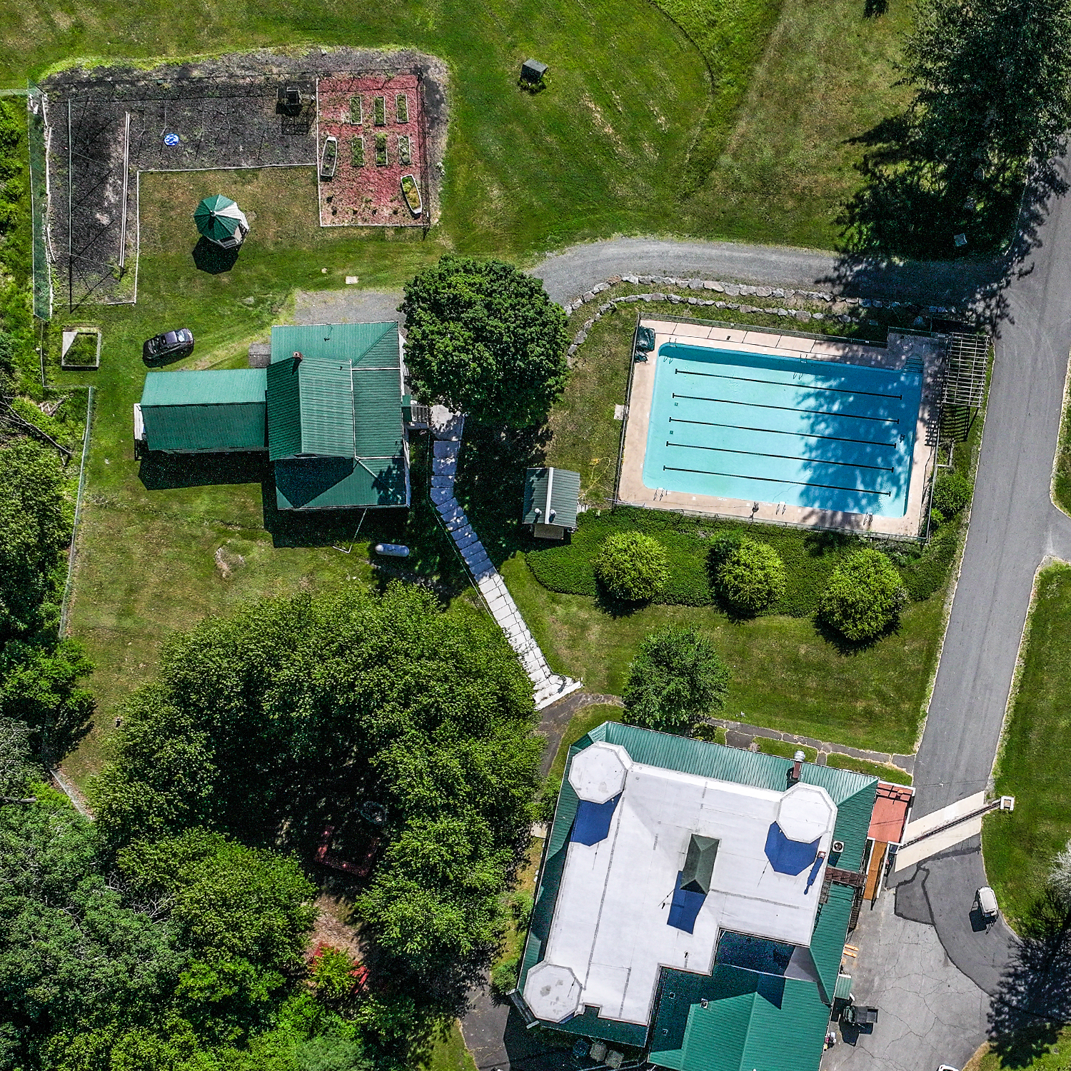 An aerial view of a backyard showing a house with a green metal roof, a swimming pool, a garden area with raised beds, a small shed, and several trees and bushes.