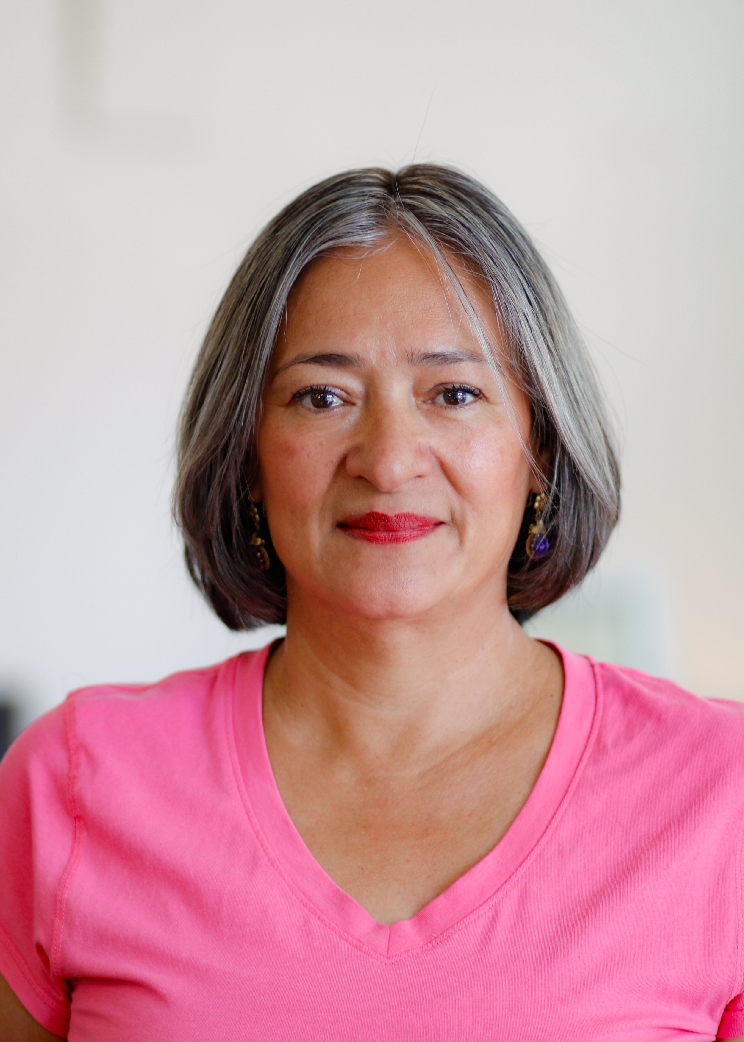 A woman with shoulder-length gray hair wearing a pink V-neck shirt and purple earrings, smiling distinctly, in an indoor setting with a blurred background.