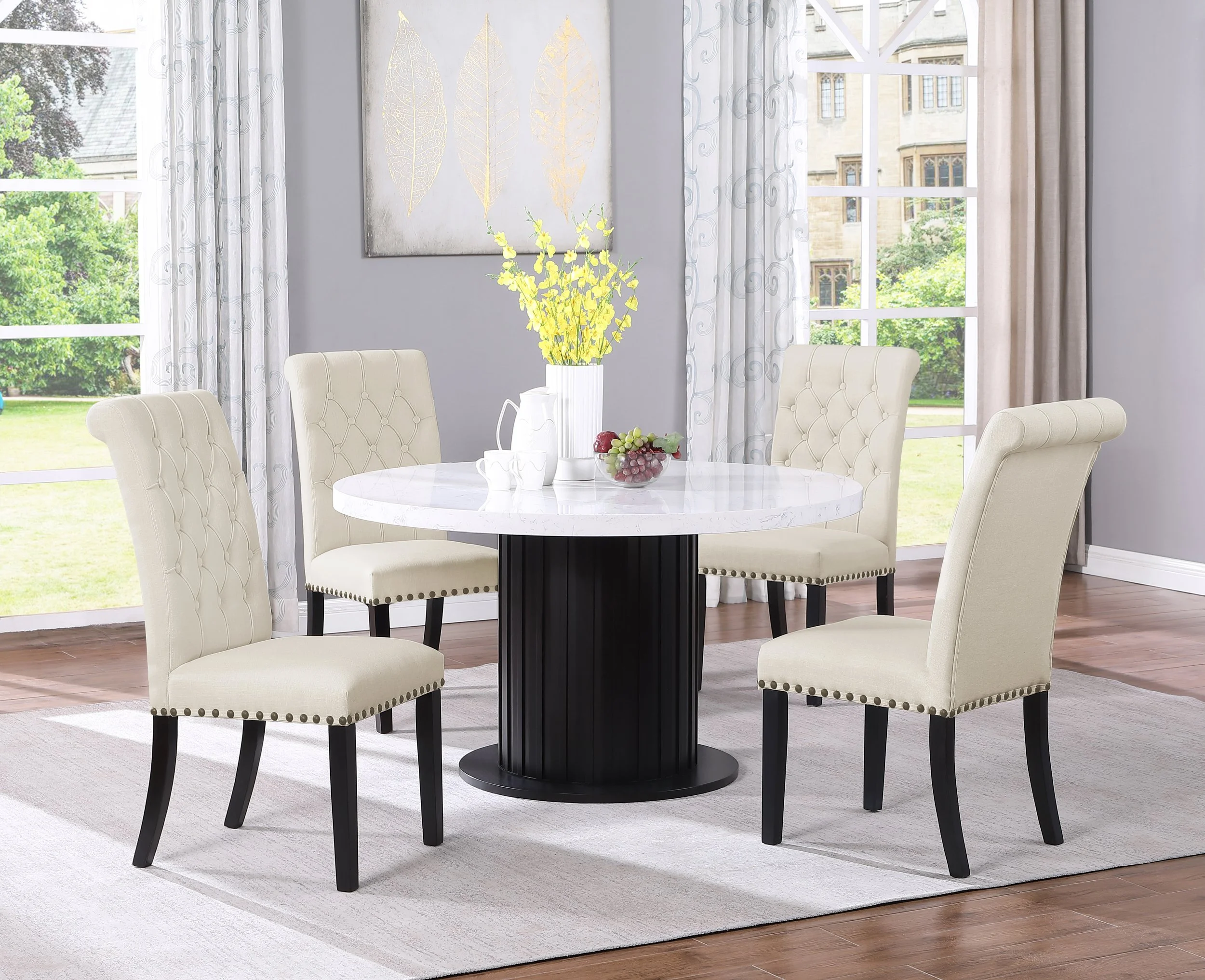 Dining room with a round white marble table, six cream tufted chairs with black legs, a white vase with yellow flowers, a bowl of grapes, and a window with white and gray curtains showing a garden outside.