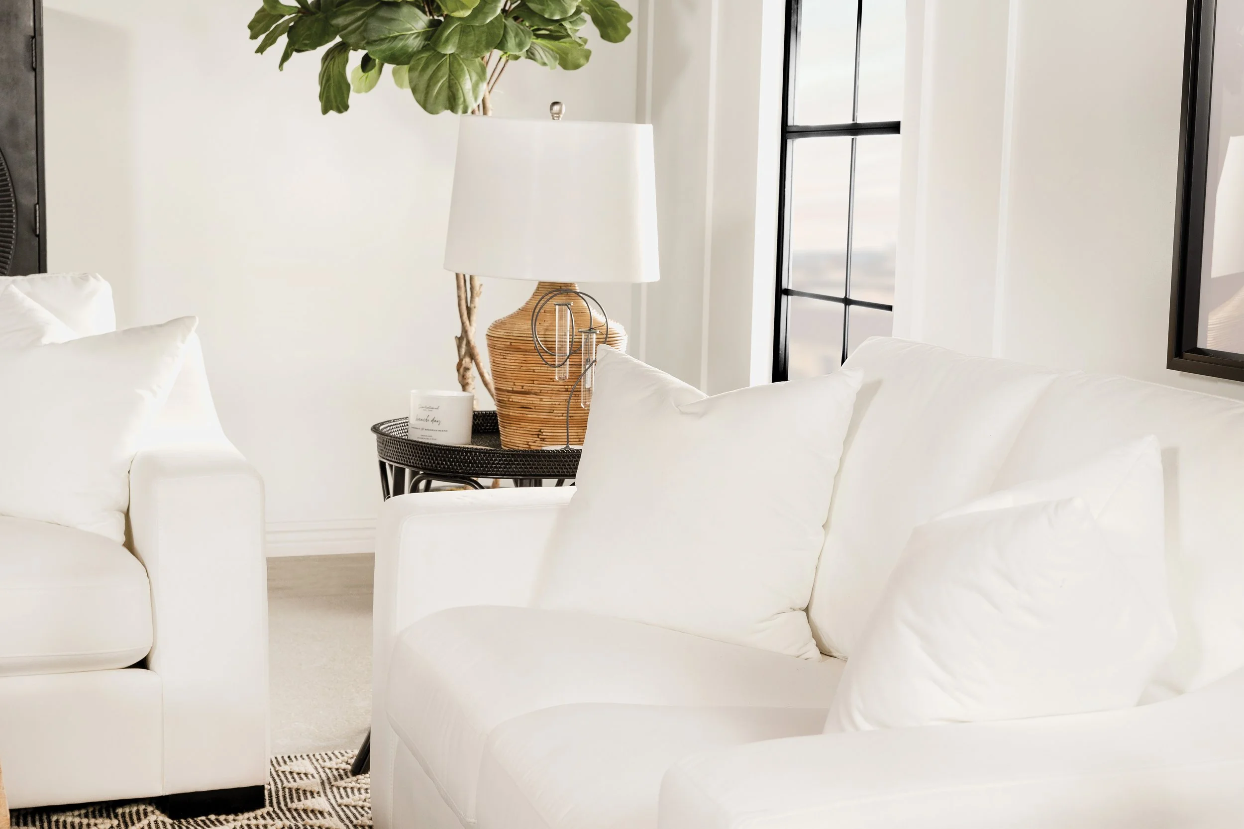 Living room corner with white sofas, black side table with lamp, large green plant, and window with black frame.