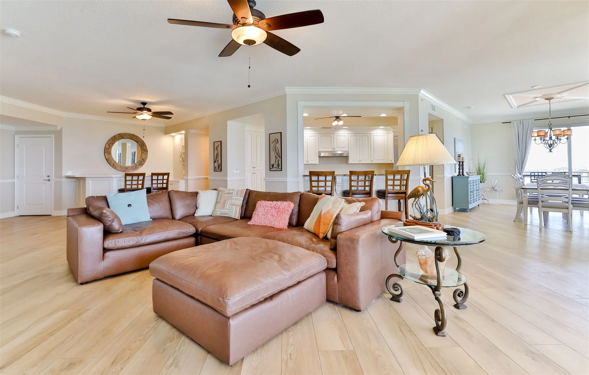 Open-concept living room with a large tan leather sectional sofa, a glass side table with a lamp, hardwood floors, ceiling fans, and a view into the kitchen and dining area with white cabinetry, a bar with chairs, and a chandelier, in a bright and ai
