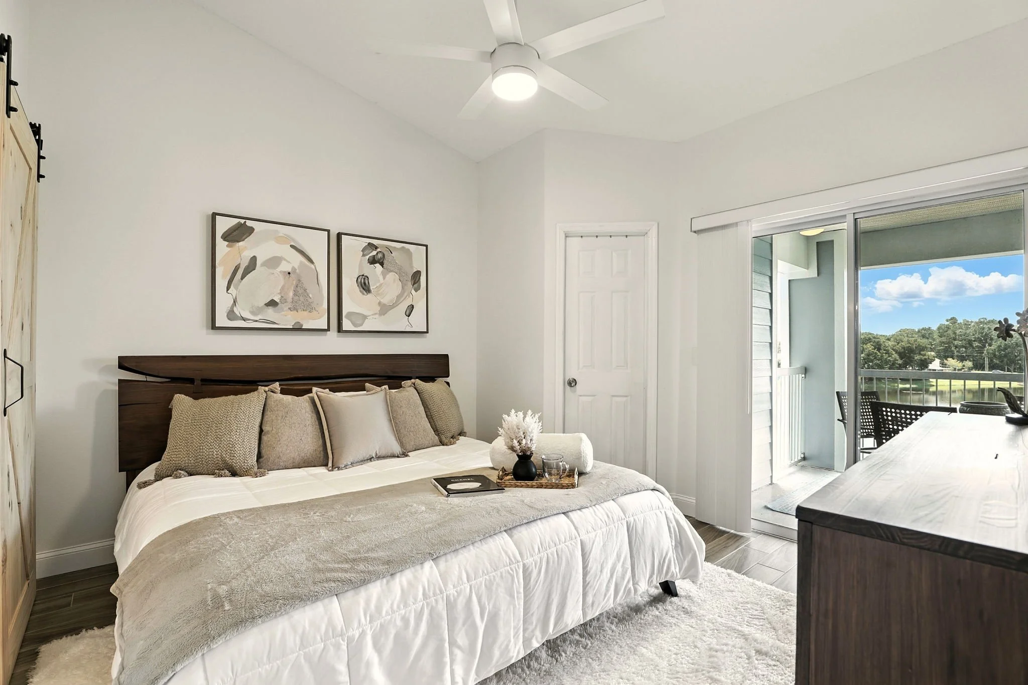 A bedroom with a bed, beige and gray pillows, wall art, a sliding glass door leading to a balcony with outdoor furniture, and a ceiling fan.