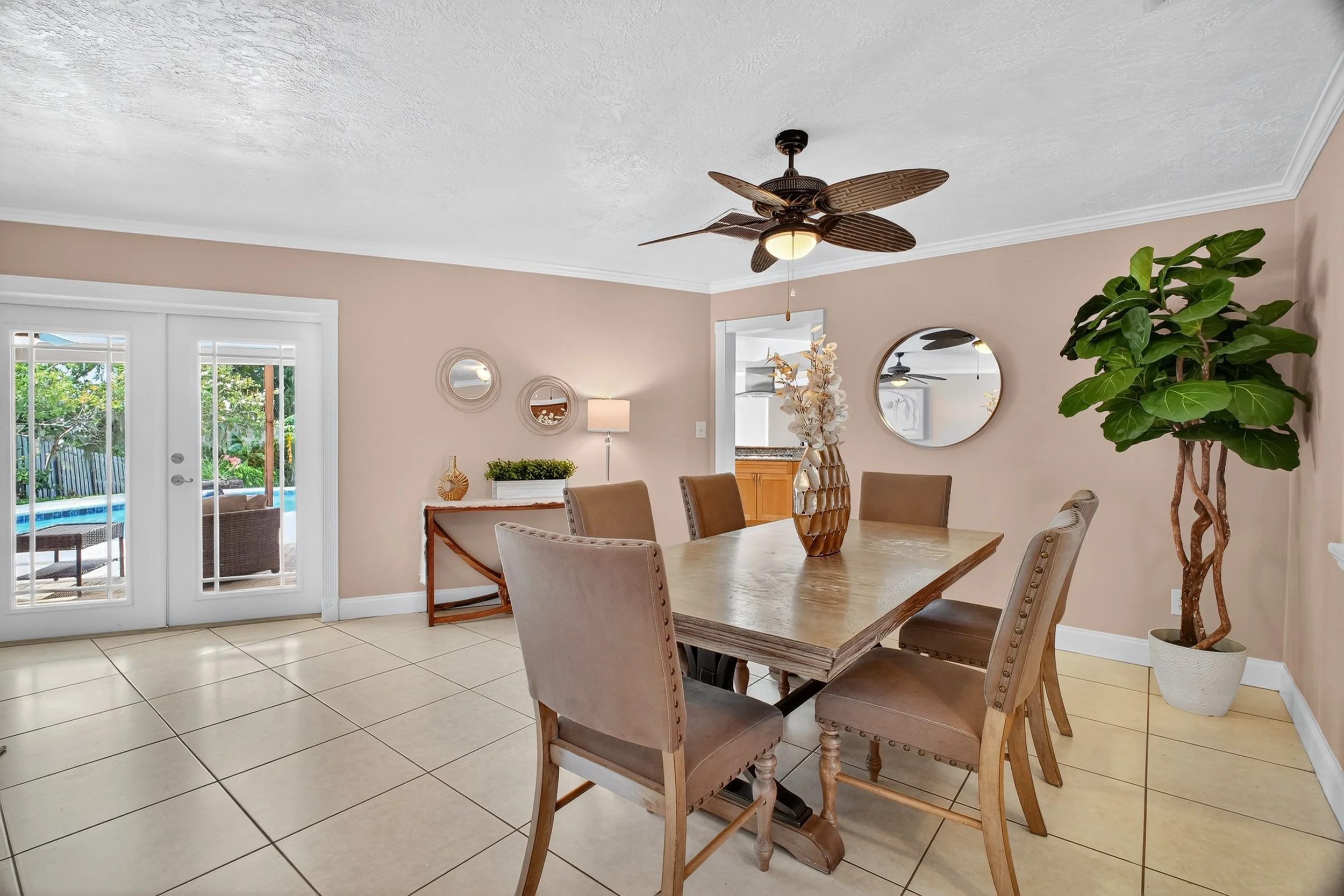 Dining room with a wooden table, six beige upholstered chairs, a ceiling fan, a potted plant, a mirror on the wall, and a sliding glass door leading outside to a backyard pool.