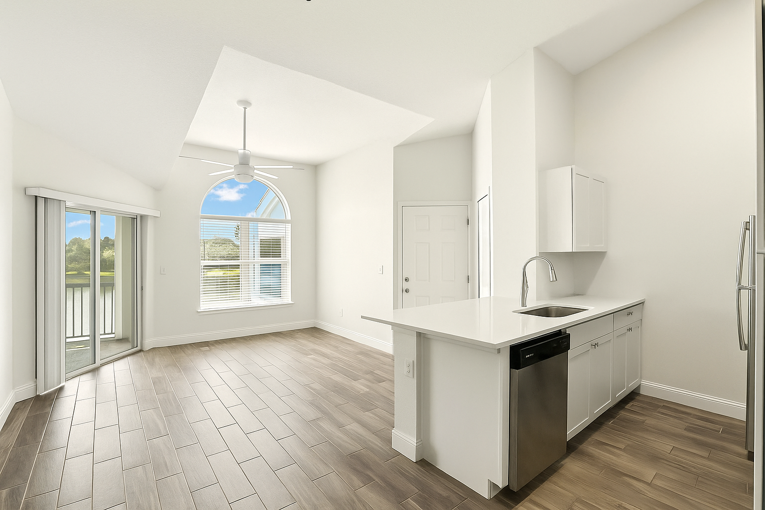 Empty kitchen and dining area with white walls, wood-like flooring, a sliding glass door leading to an outdoor space, a large arched window, a ceiling fan, and kitchen cabinets with a sink and dishwasher.