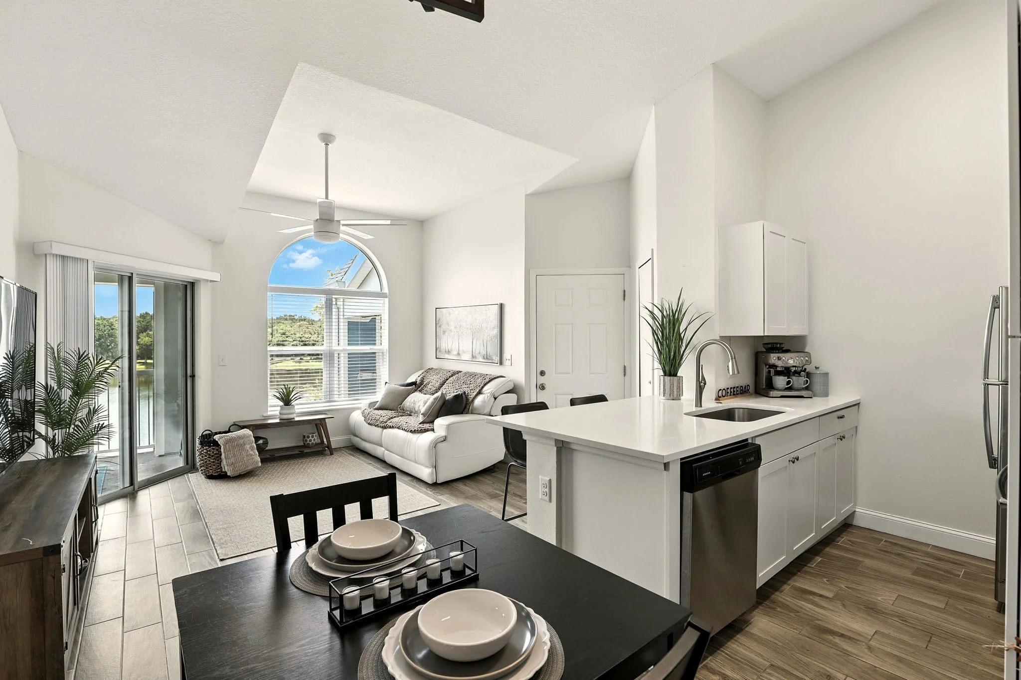 Open-concept living room and kitchen with white walls, hardwood flooring, a white couch, a black dining table with place settings, a sliding glass door, and a large window showing a view of trees and a blue sky.