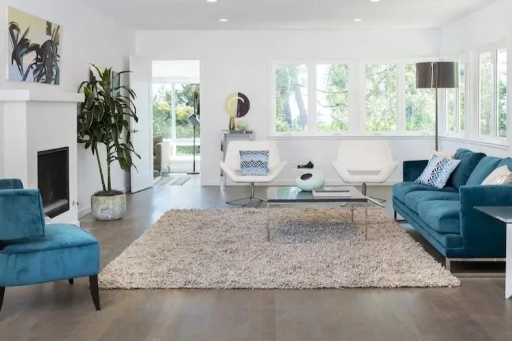 Bright living room with white walls, large windows, and modern furniture including a blue sofa, white armchair, and a glass coffee table on a plush beige rug. Decoraive items and plants are also visible.