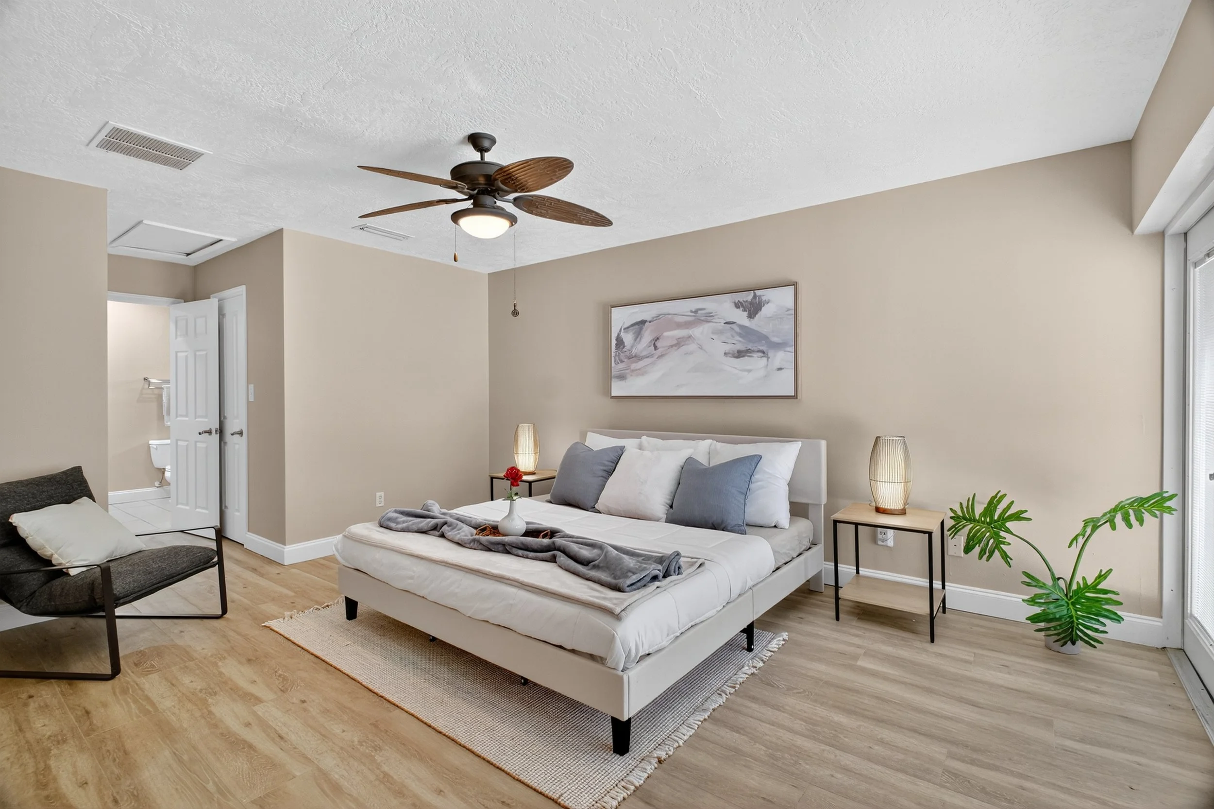 A bedroom with beige walls, light wood flooring, a white bed with white and gray pillows, and a gray throw blanket. There are two bedside tables with lamps, a wall art above the bed, a black armchair with a white pillow, a potted plant, and a sliding