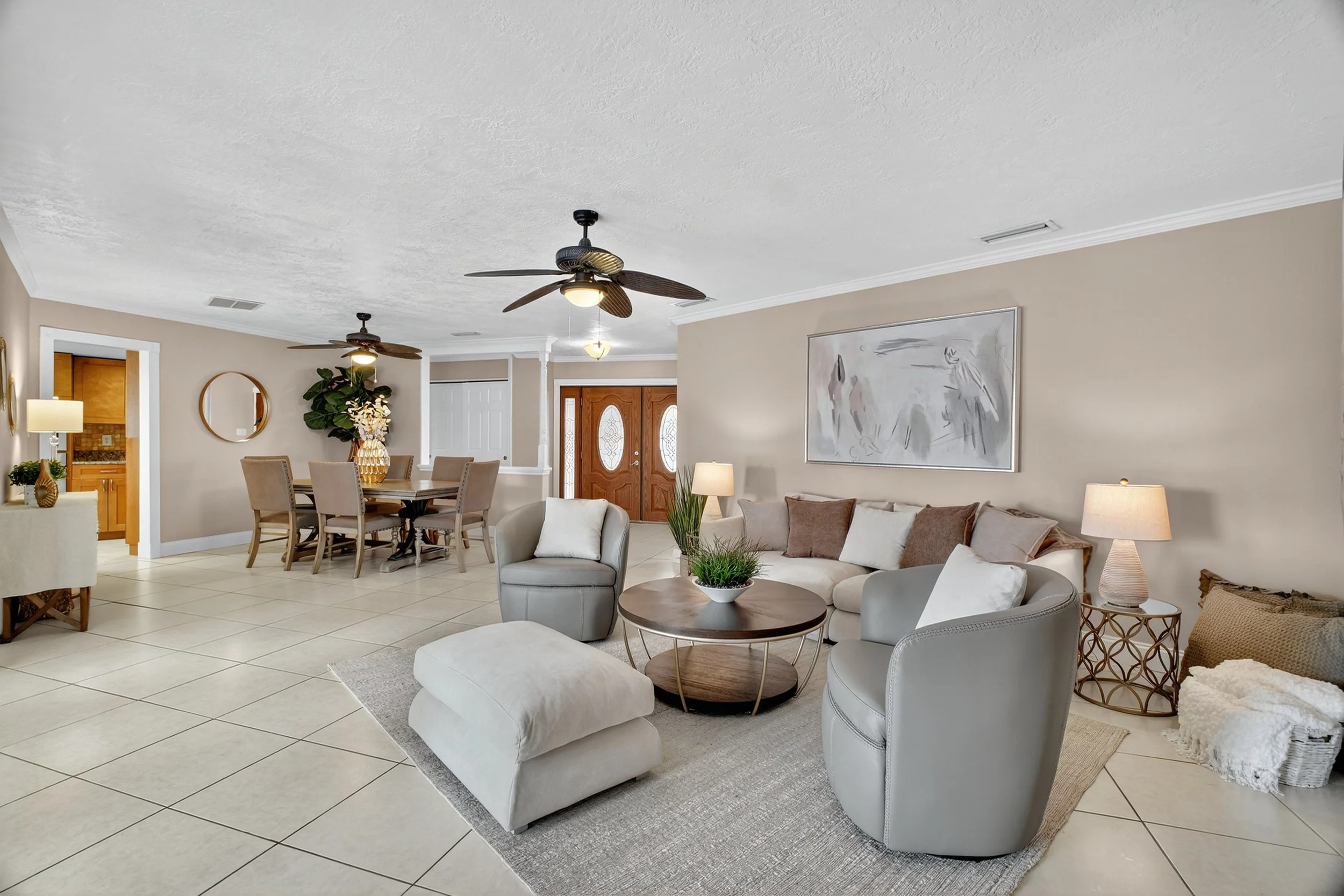 Spacious living room with beige walls, white ceiling, tiled floor, beige sofa, two white armchairs, gray ottoman, wooden coffee table with plants, two side lamps, abstract wall art, and a dining area with a wooden table and six chairs.