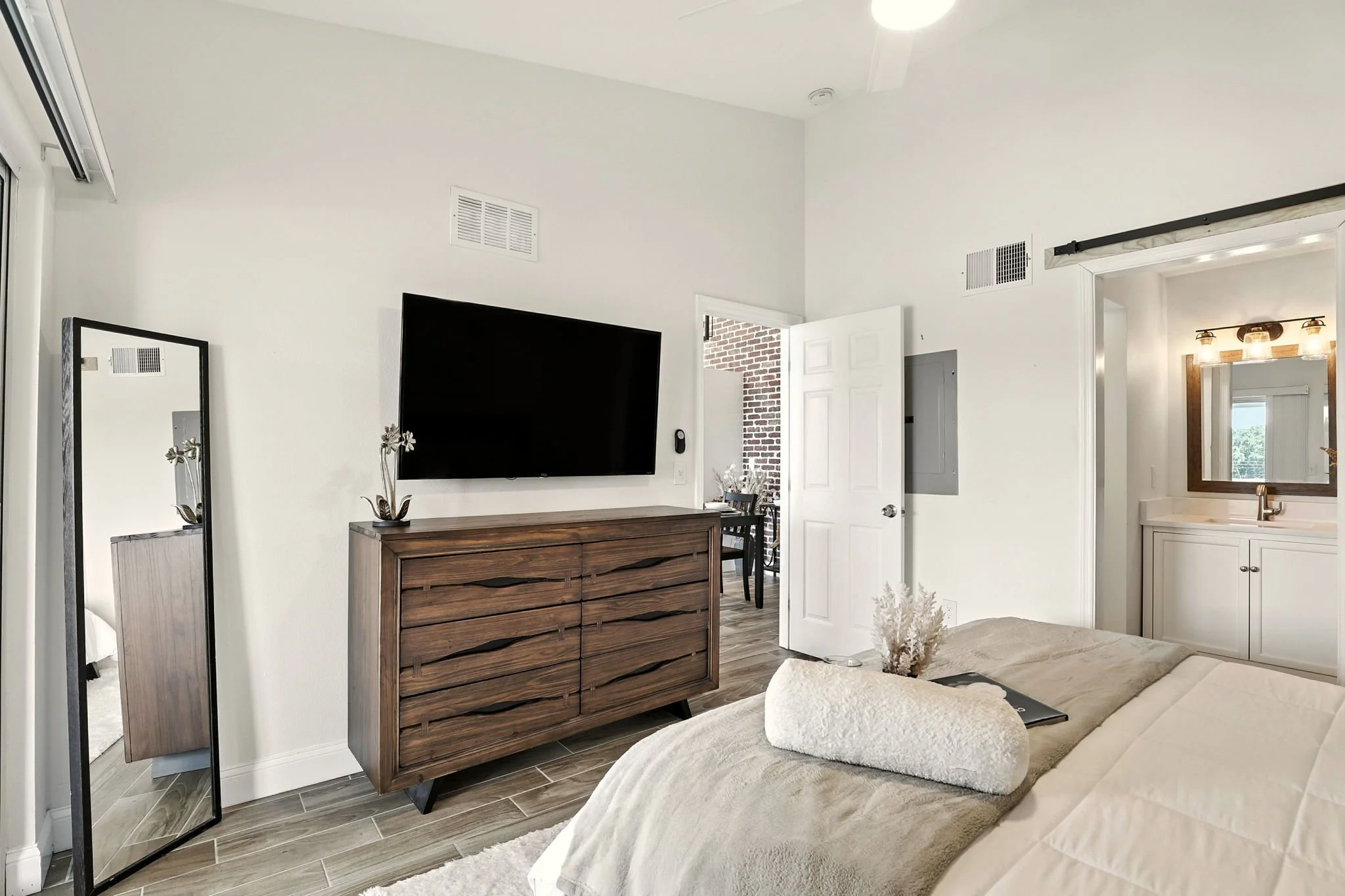 Modern bedroom with a on-wall TV, leaning mirror, wooden dresser, bed with beige bedding, and an attached bathroom with mirror and vanity.