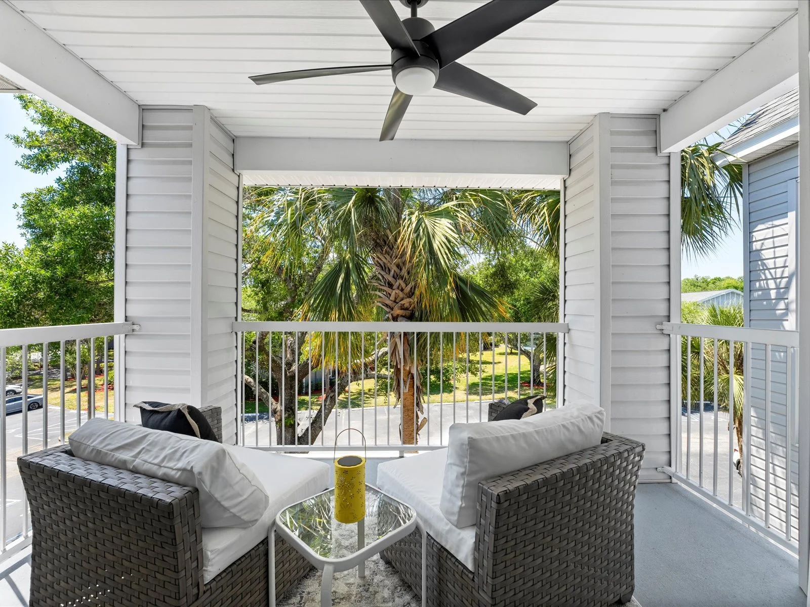 A balcony with two wicker chairs and white cushions, a small glass-top table with a yellow candle holder, overlooking green trees including a large palm tree, with a white railing and siding, under a ceiling fan.