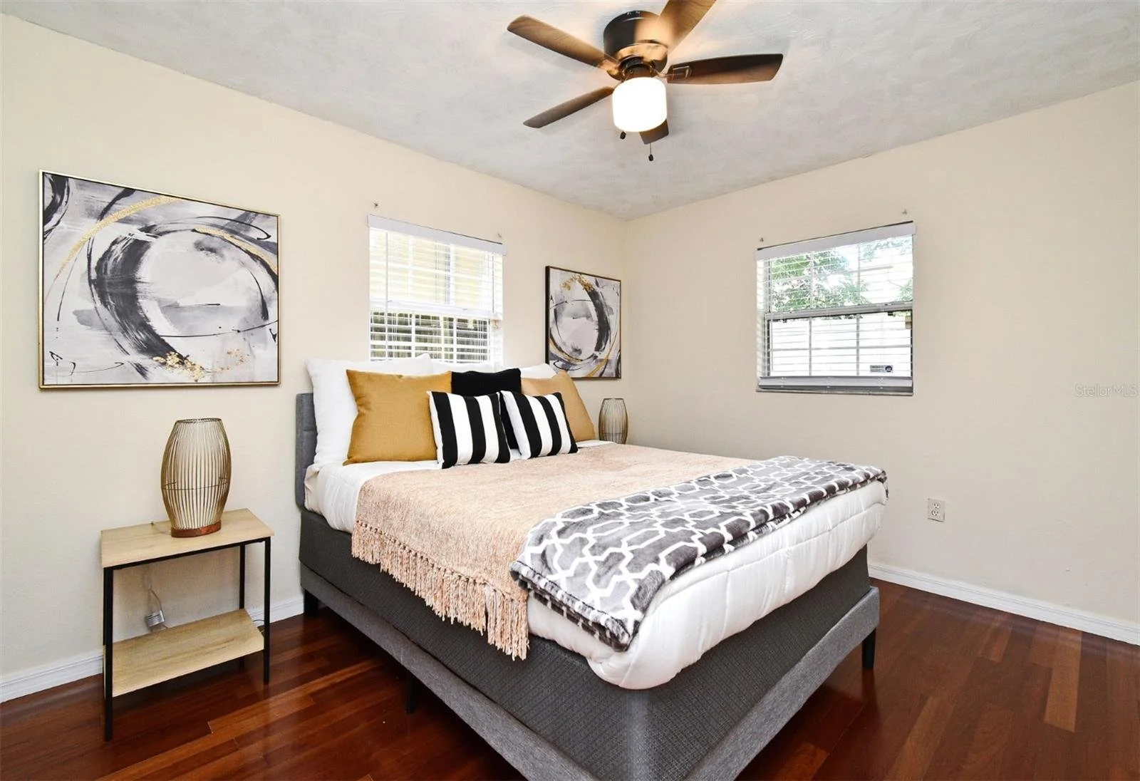A bedroom with a gray upholstered bed, beige pillows, black and white striped pillow, and blanket. There are two abstract black, gray, and gold paintings on the powder-colored walls, accompanied by a wooden nightstand with a tall decorative lamp, pol