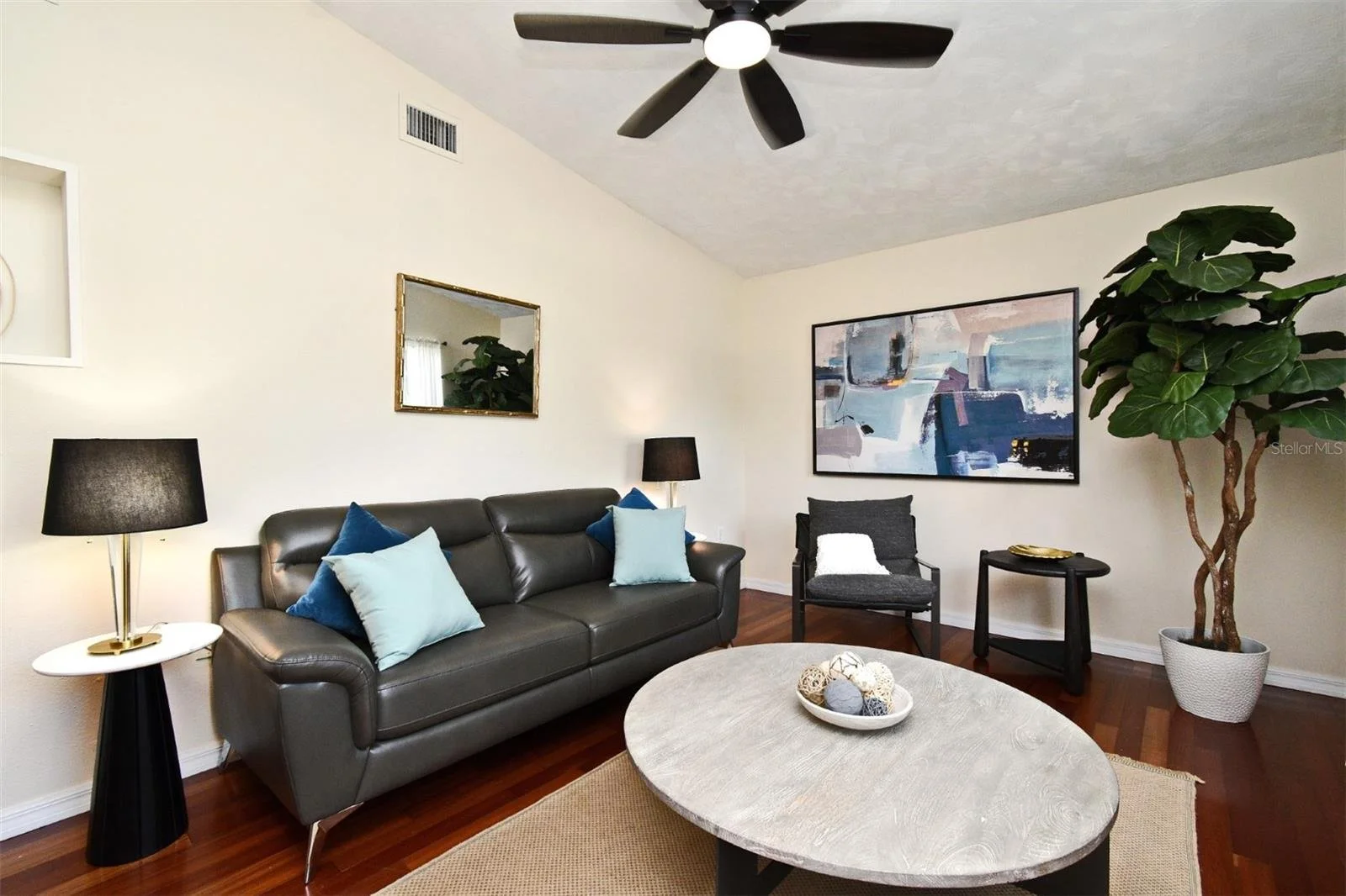 Living room with a dark leather sofa adorned with blue and light blue throw pillows, black table lamps, a large abstract painting on the wall, a potted fiddle leaf fig tree, a round coffee table decorated with a bowl of decorative balls, and wooden f