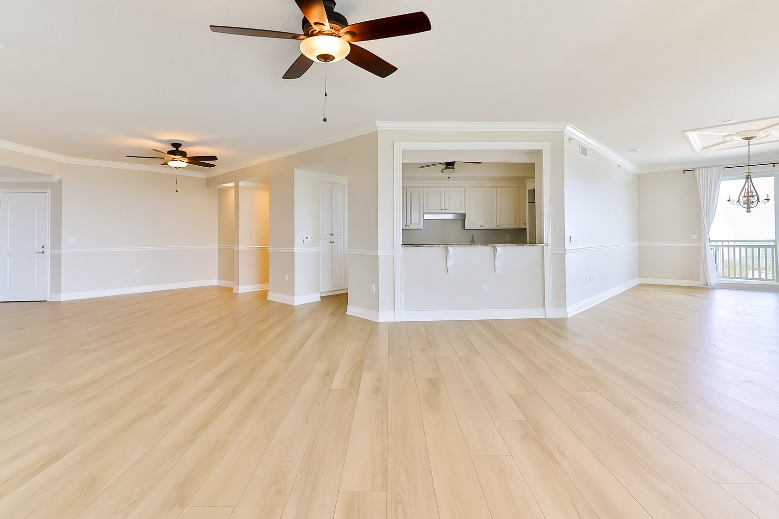 Spacious, empty living room with light wood flooring, white walls, ceiling fans, large window with curtains, and an open kitchen area.
