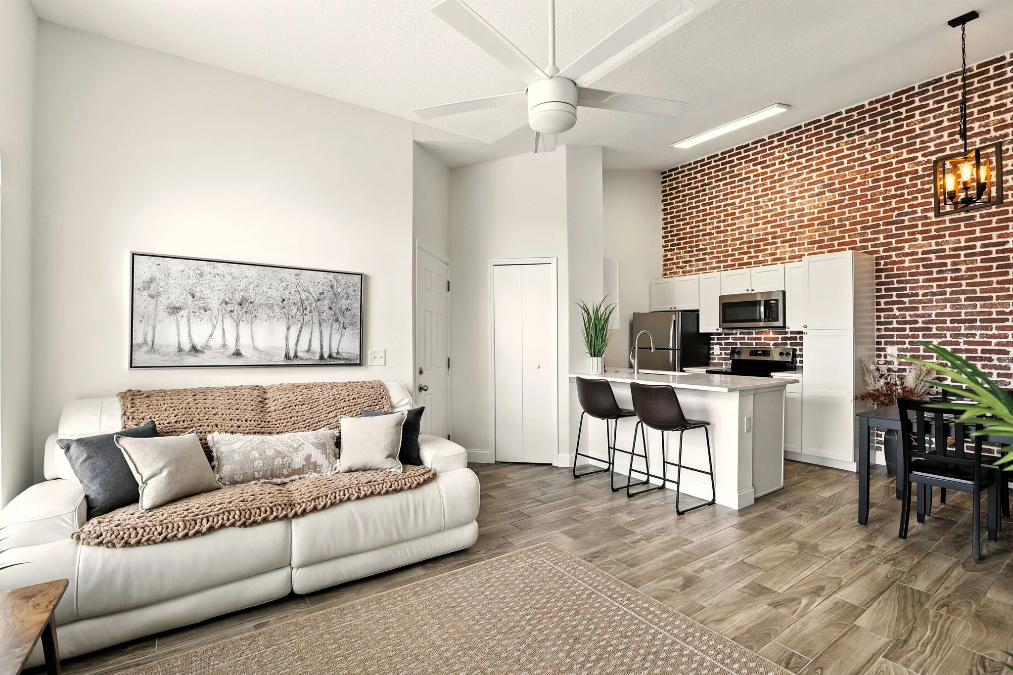 Open-concept living room and kitchen with white walls, wood flooring, a white sofa with cushions, a black and white artwork above the sofa, a ceiling fan, brick accent wall, white cabinets, a stainless steel refrigerator, microwave, and oven, two bla
