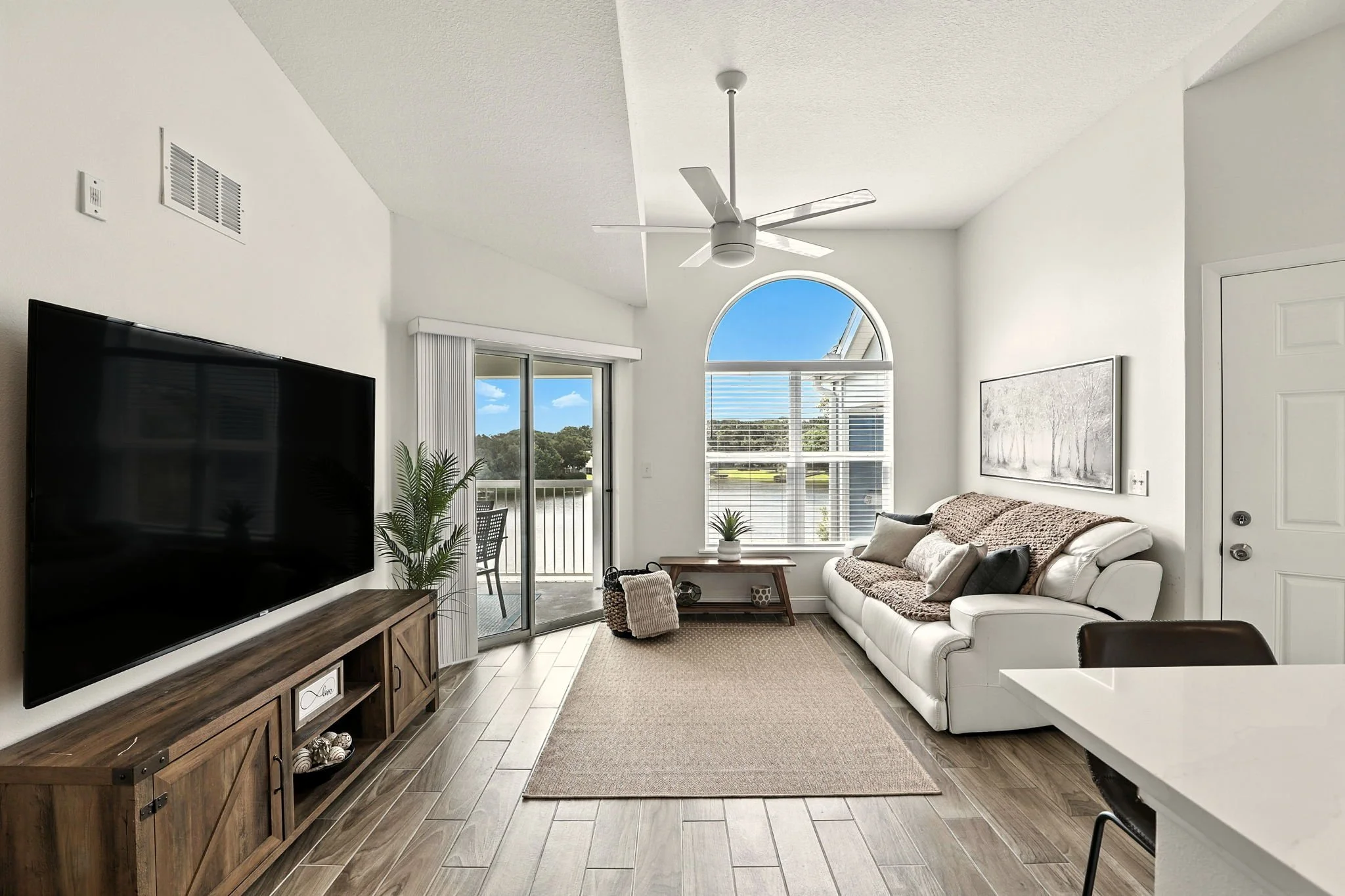 Living room with white walls, wooden floor, large flat-screen TV on a wooden console, white sofa with pillows, glass sliding door leading to a balcony, large window with blinds, ceiling fan, and a coffee table.